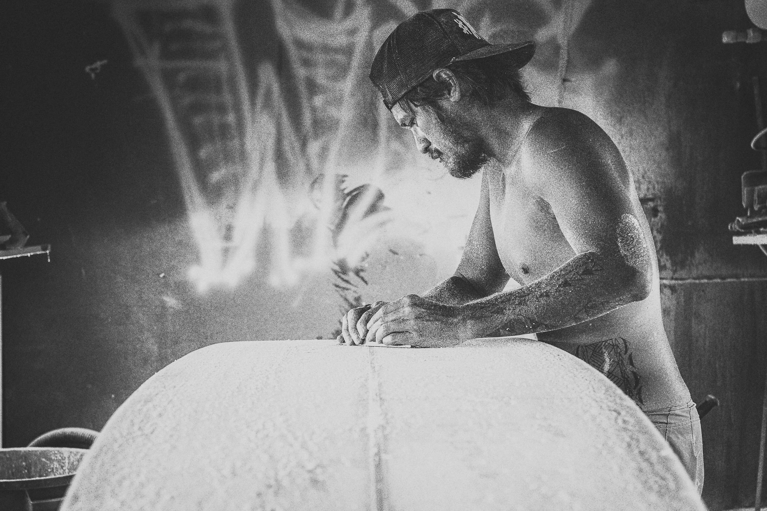 A man working on a surfboard in a workshop, wearing a cap and no shirt, with a focused expression.
