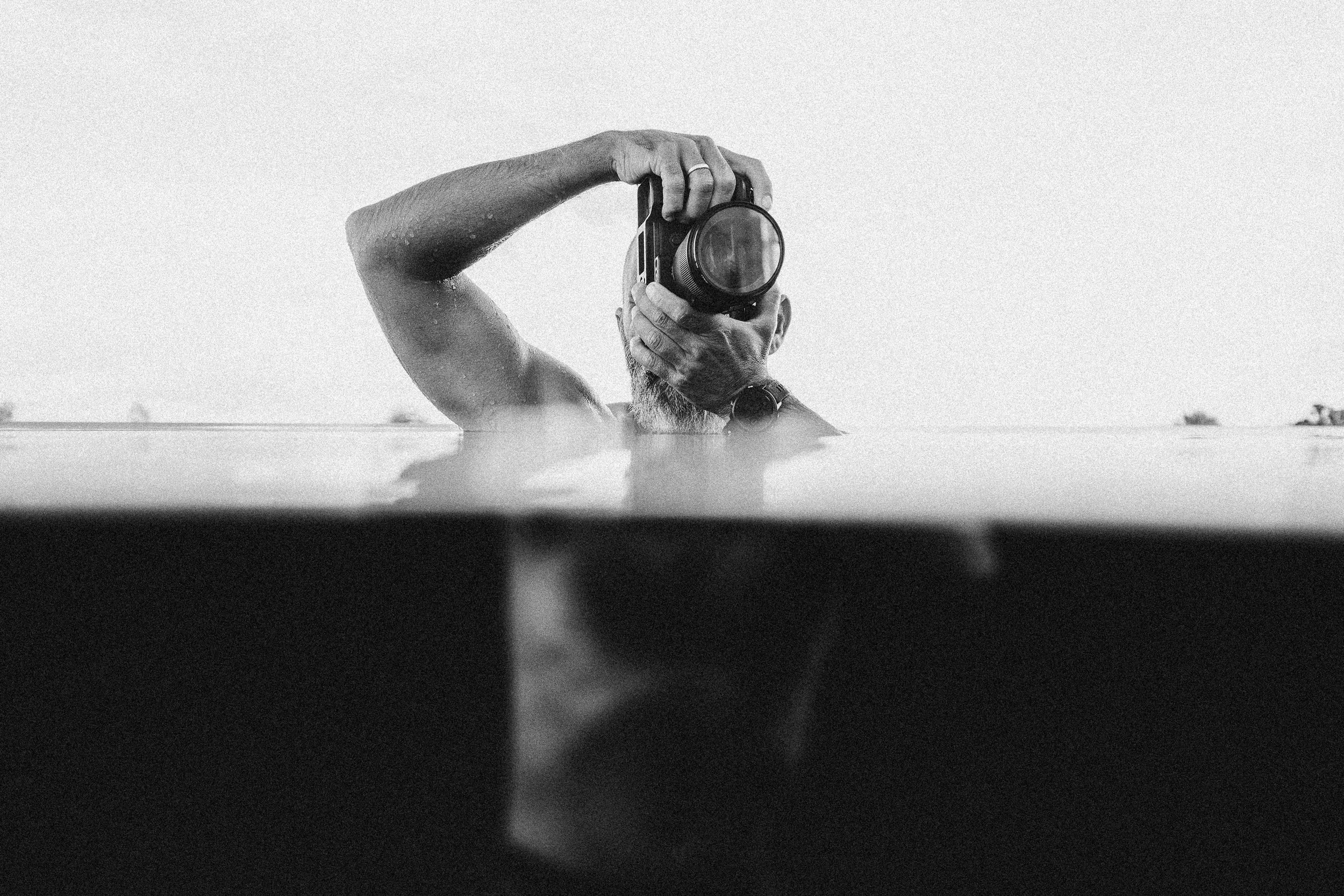 A person holding a camera above their head, partially submerged in water, with a watch on their wrist and water droplets on their arm, in black and white.