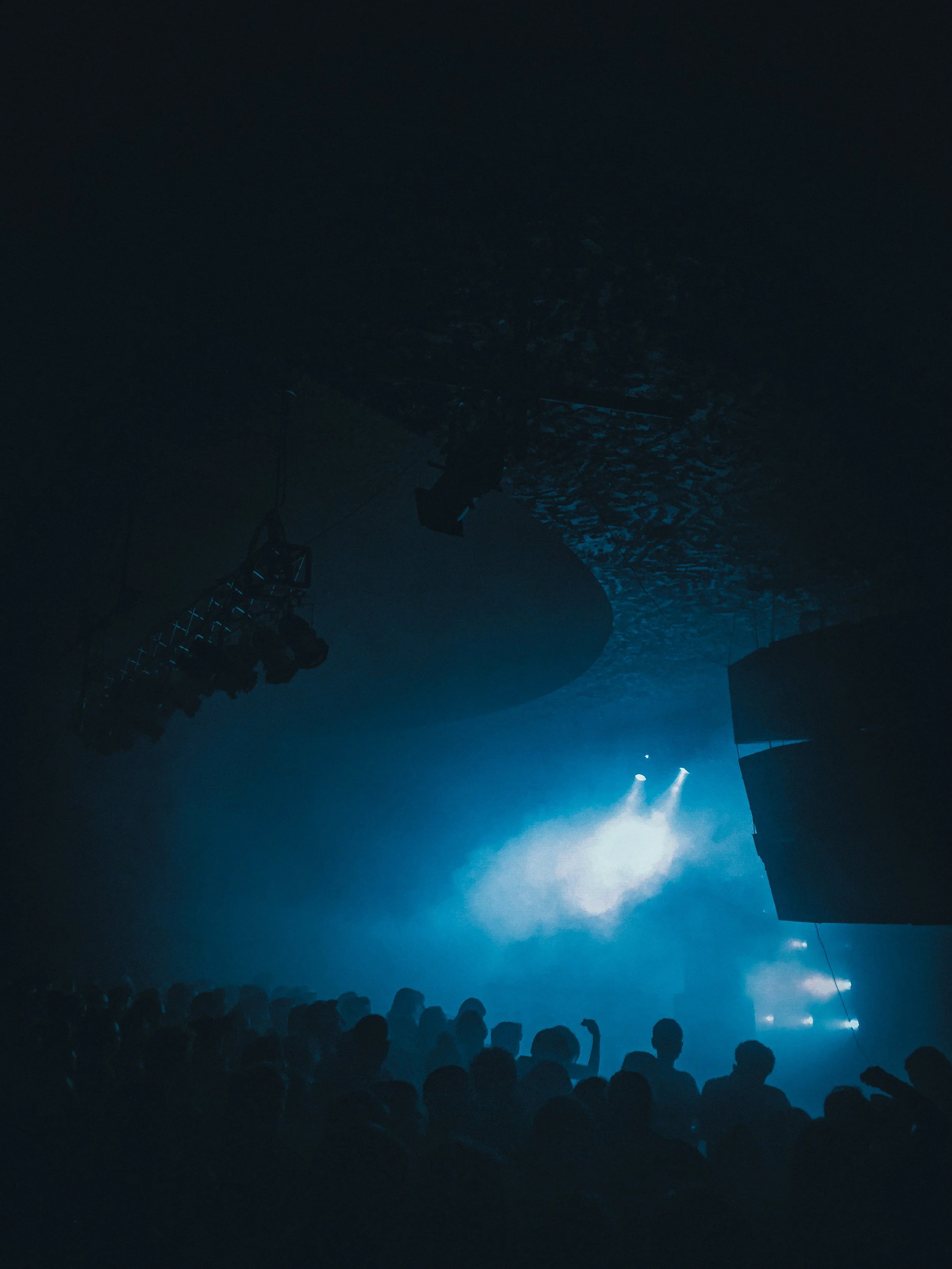 People attending a concert or performance in a dark venue with blue lighting, audience silhouette visible at the bottom, stage and equipment overhead.