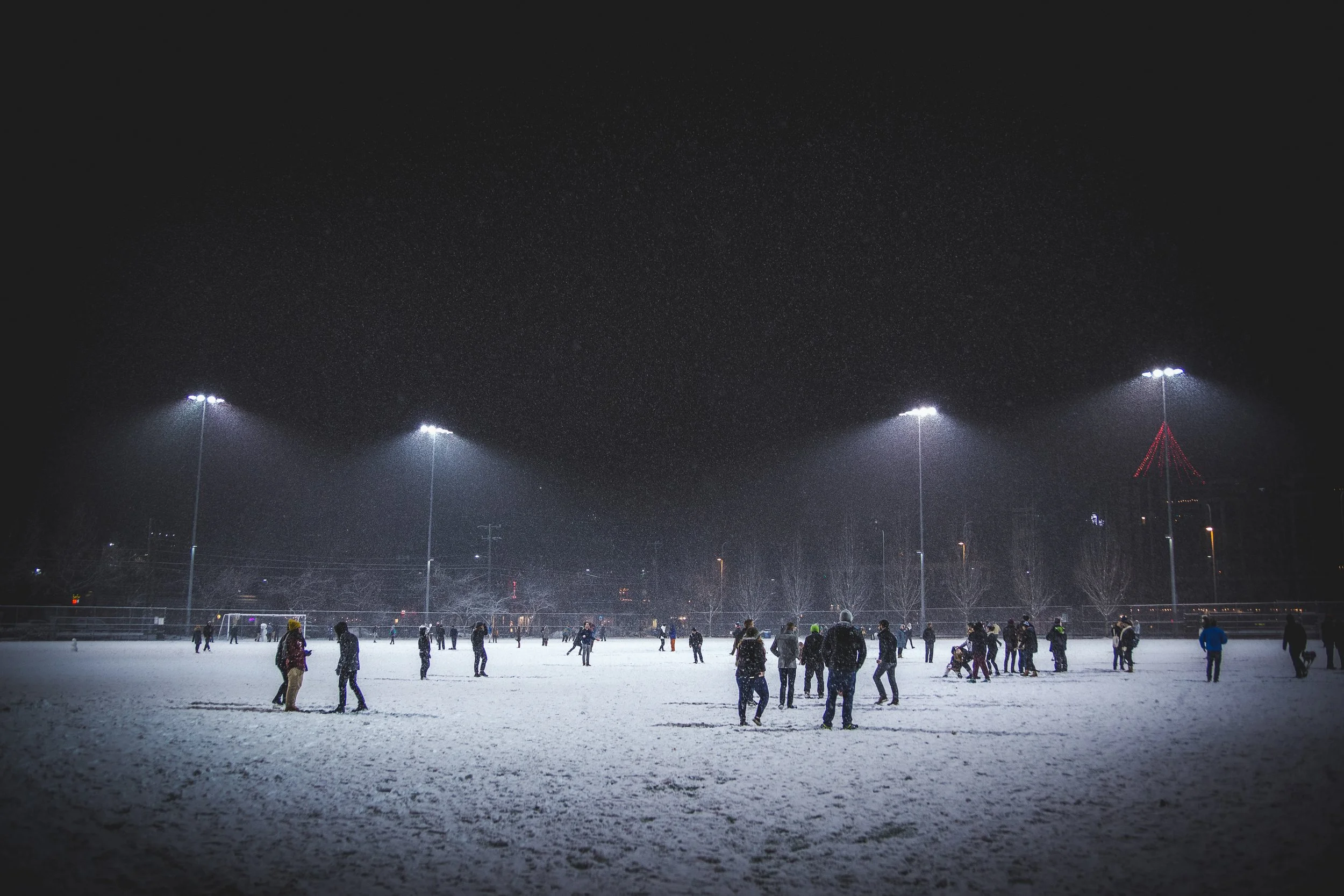 People ice skating at night on an outdoor ice rink under bright overhead lights, with a dark sky overhead and a distant illuminated tower.