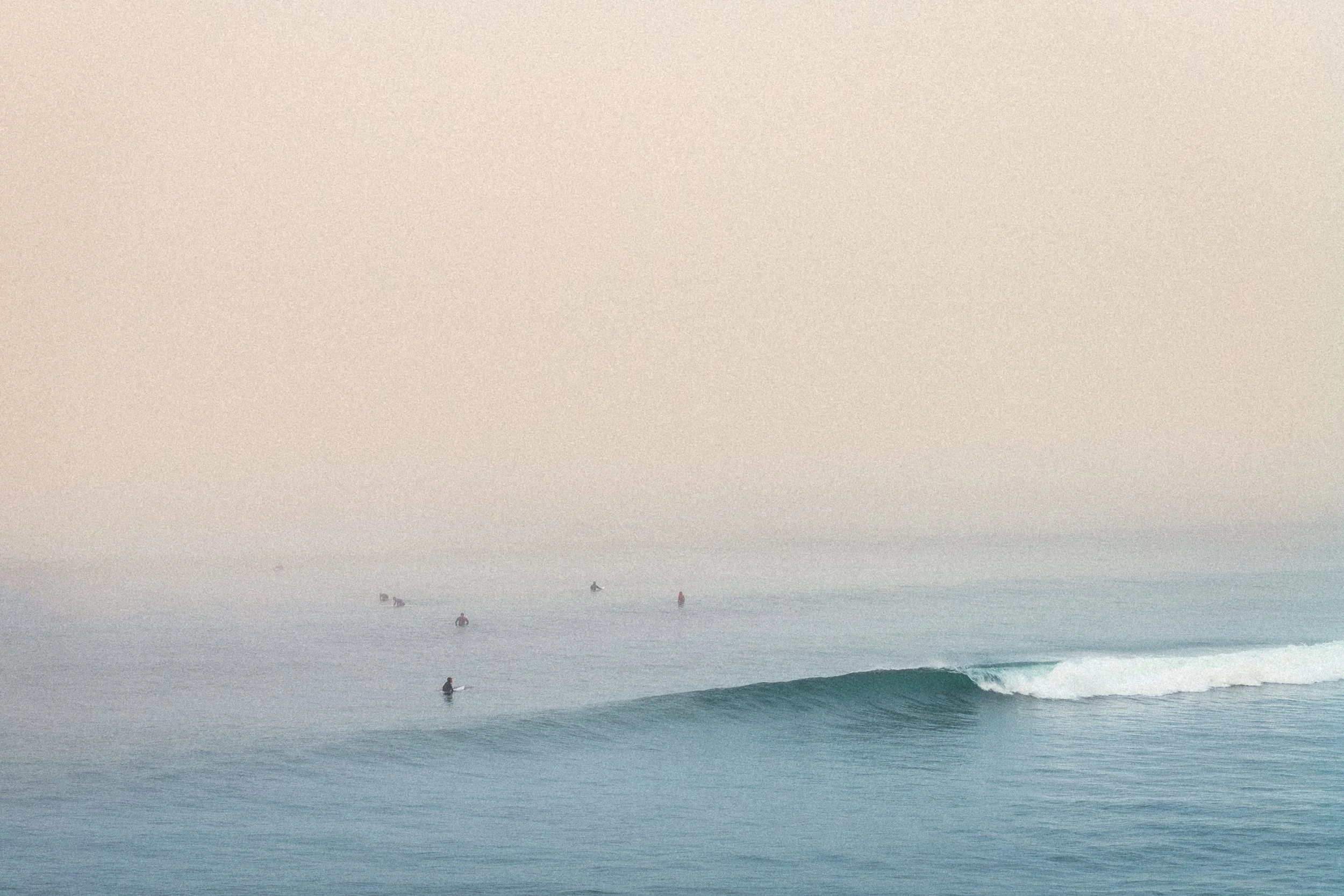 A foggy day at the beach with several people in the water and on surfboards, with ocean waves in the foreground.
