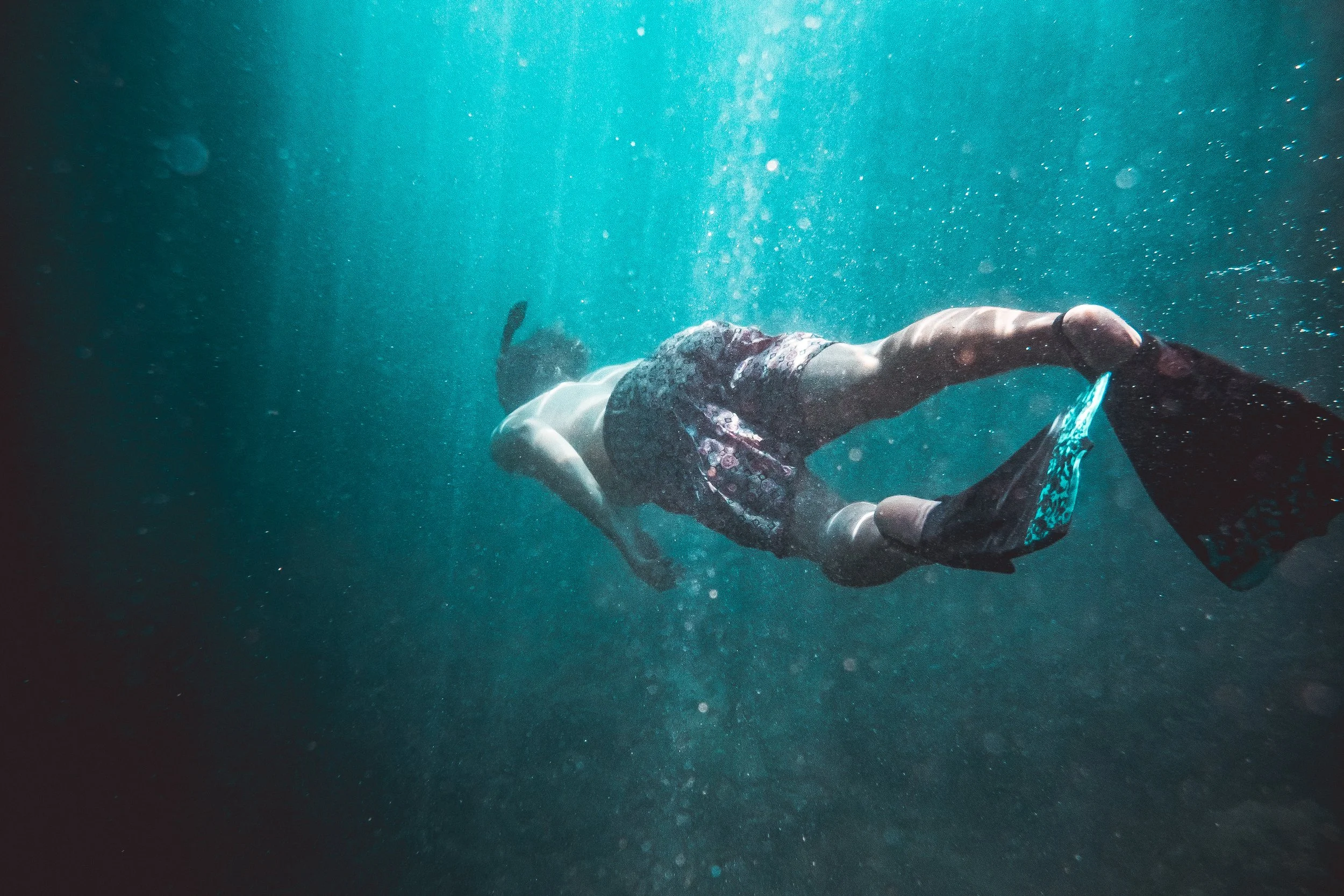 A person swimming underwater wearing patterned swim trunks, with their back to the camera and their arms extended forward.