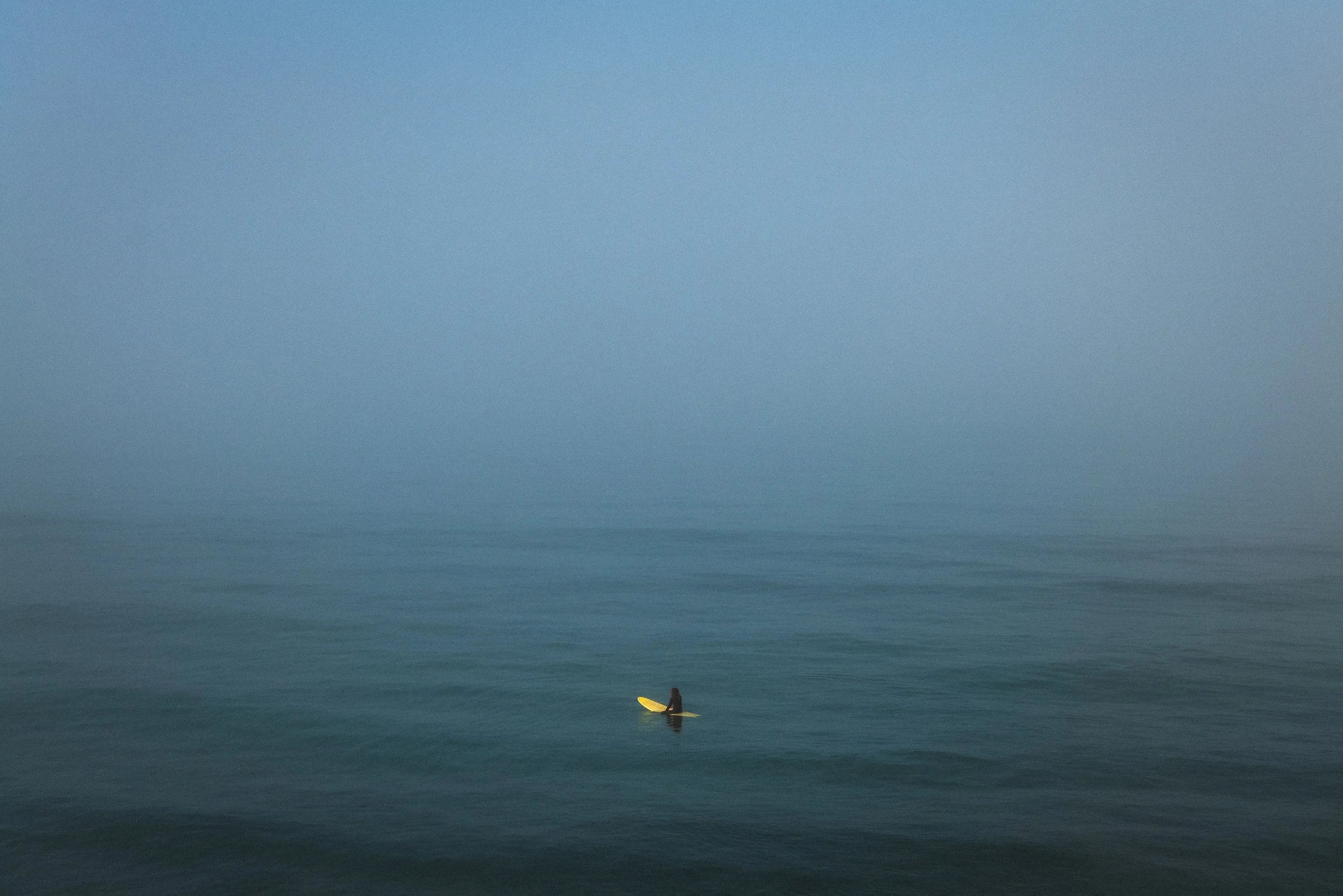 A person with a yellow surfboard sitting on a small wave in the ocean, with a calm and foggy atmosphere.