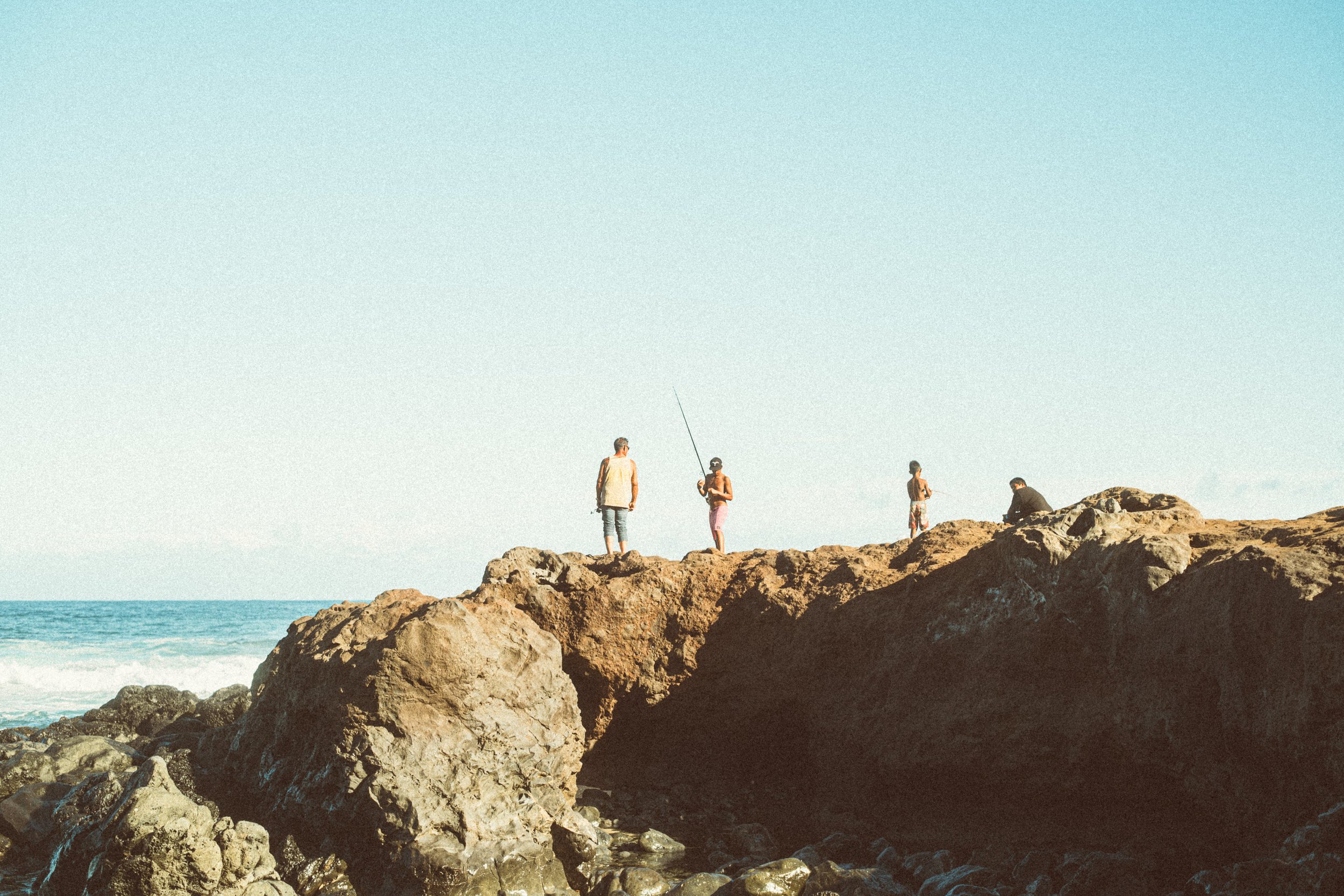 People standing and sitting on rocky terrain near the ocean, with a clear sky above.