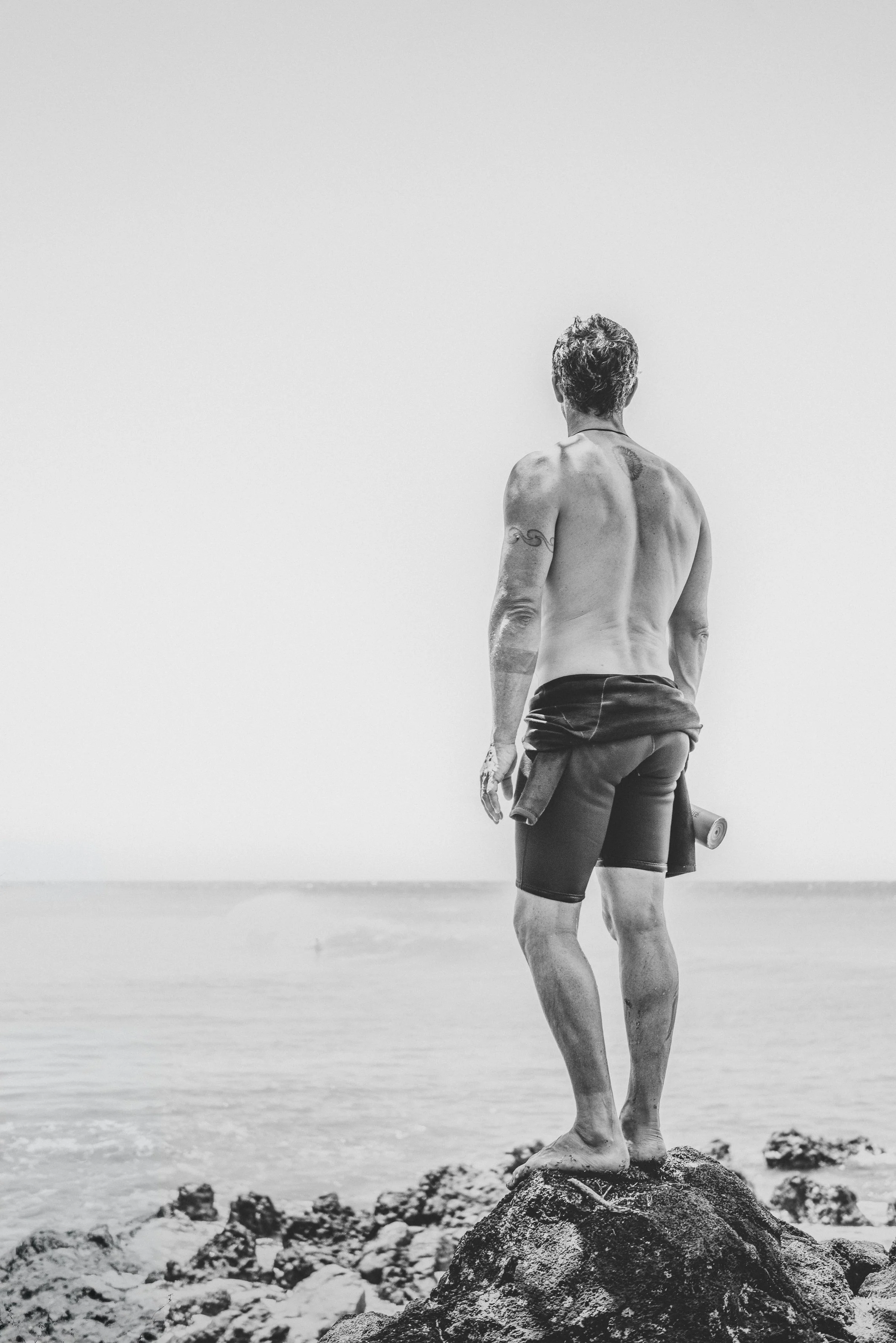 A person with short curly hair, tattoos on their arm, and wearing shorts and a jacket around their waist, stands on a rock at the beach facing away from the camera. The person appears to be practicing yoga or meditation, looking out toward the ocean 