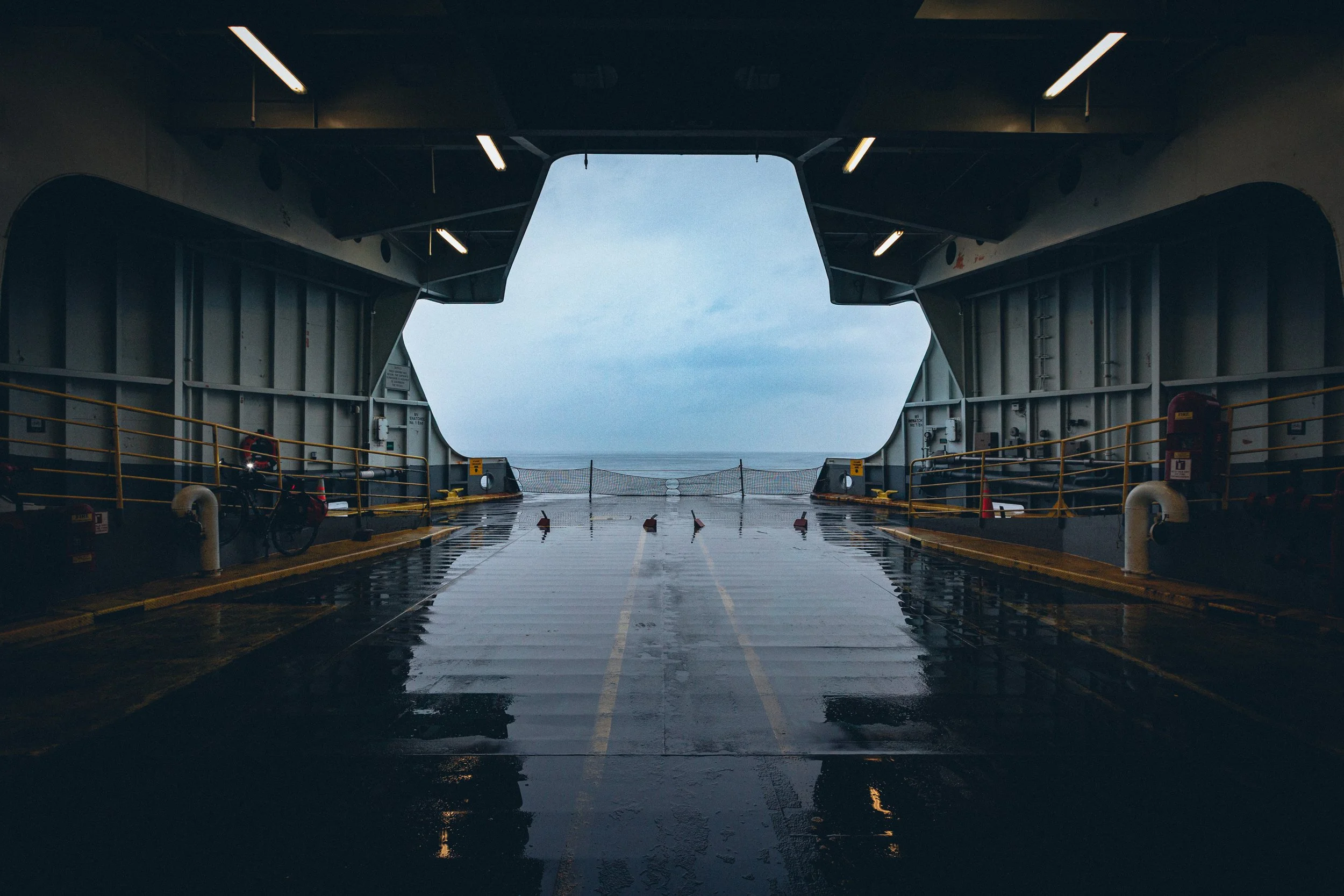 View from inside a ship's hangar or cargo bay looking out to the ocean through an open doorway, with wet reflective floors and safety railings.