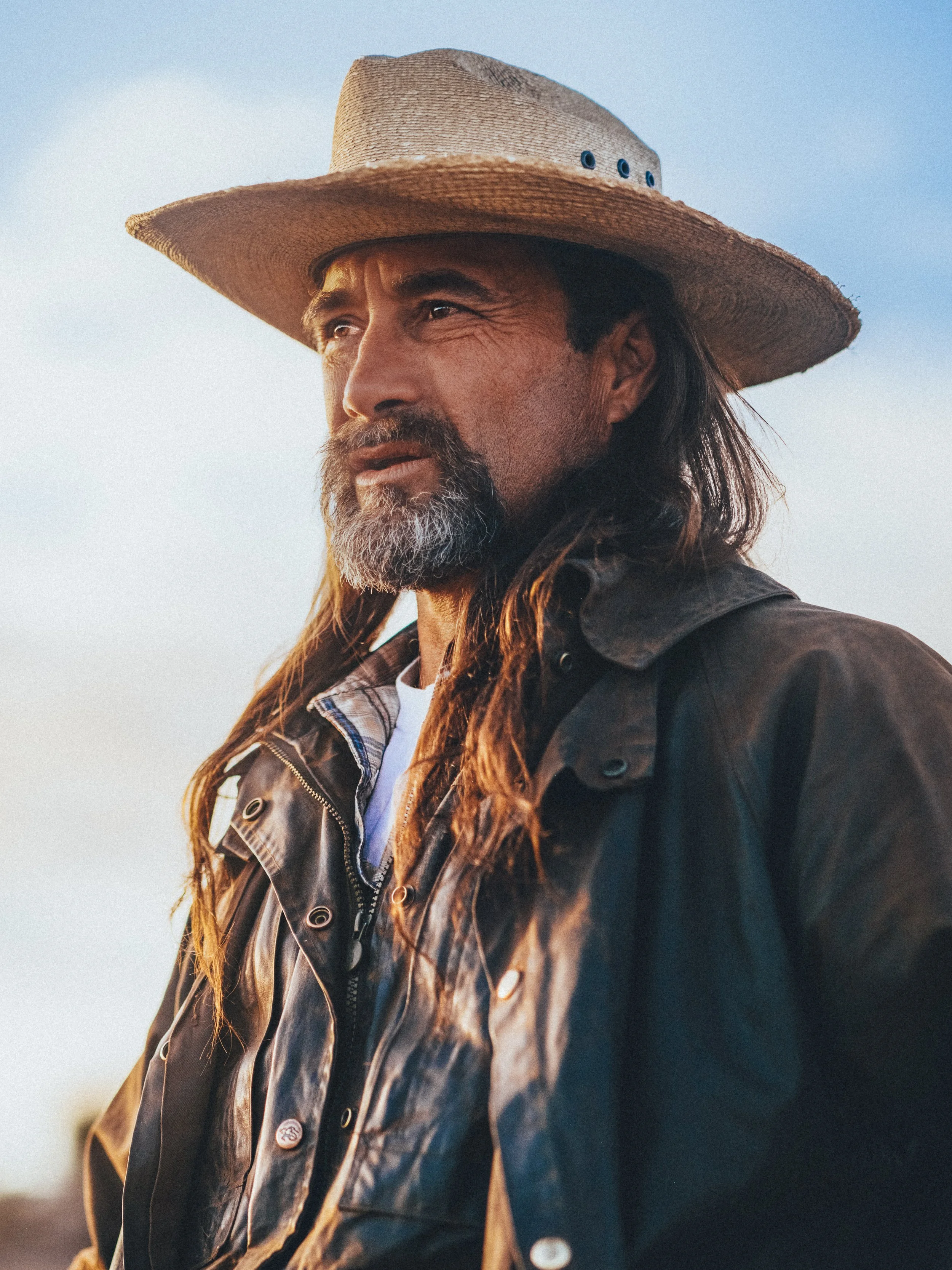 Close-up of a man with long hair and a beard wearing a wide-brimmed straw hat and a dark jacket, outdoors during the daytime.