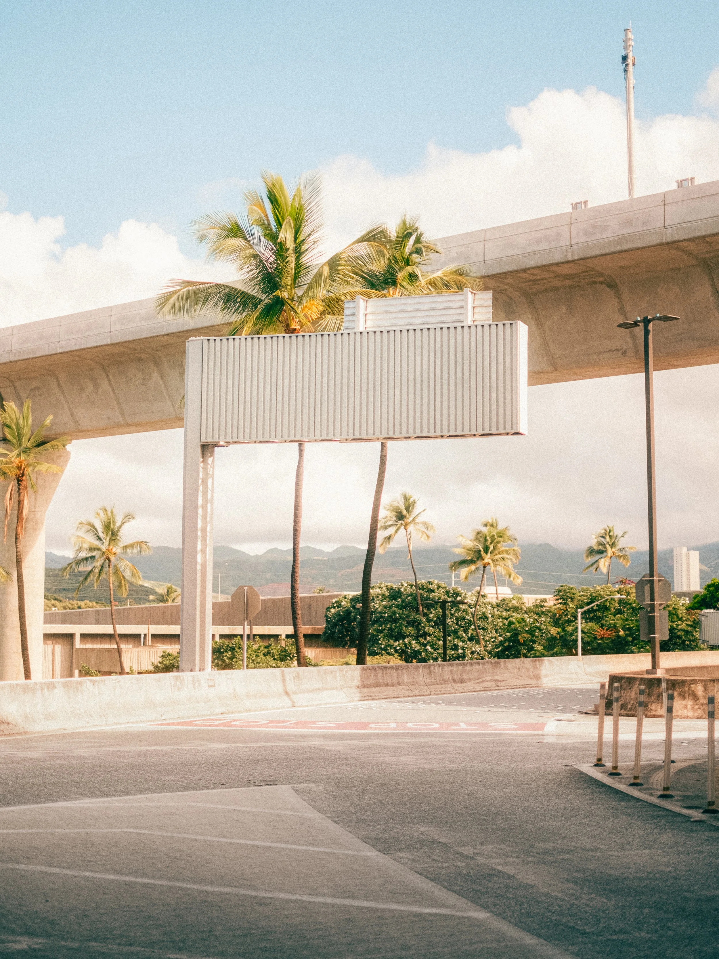 Empty parking lot beneath a bridge with palm trees in the background, cloudy sky, and a large billboard frame without signage.