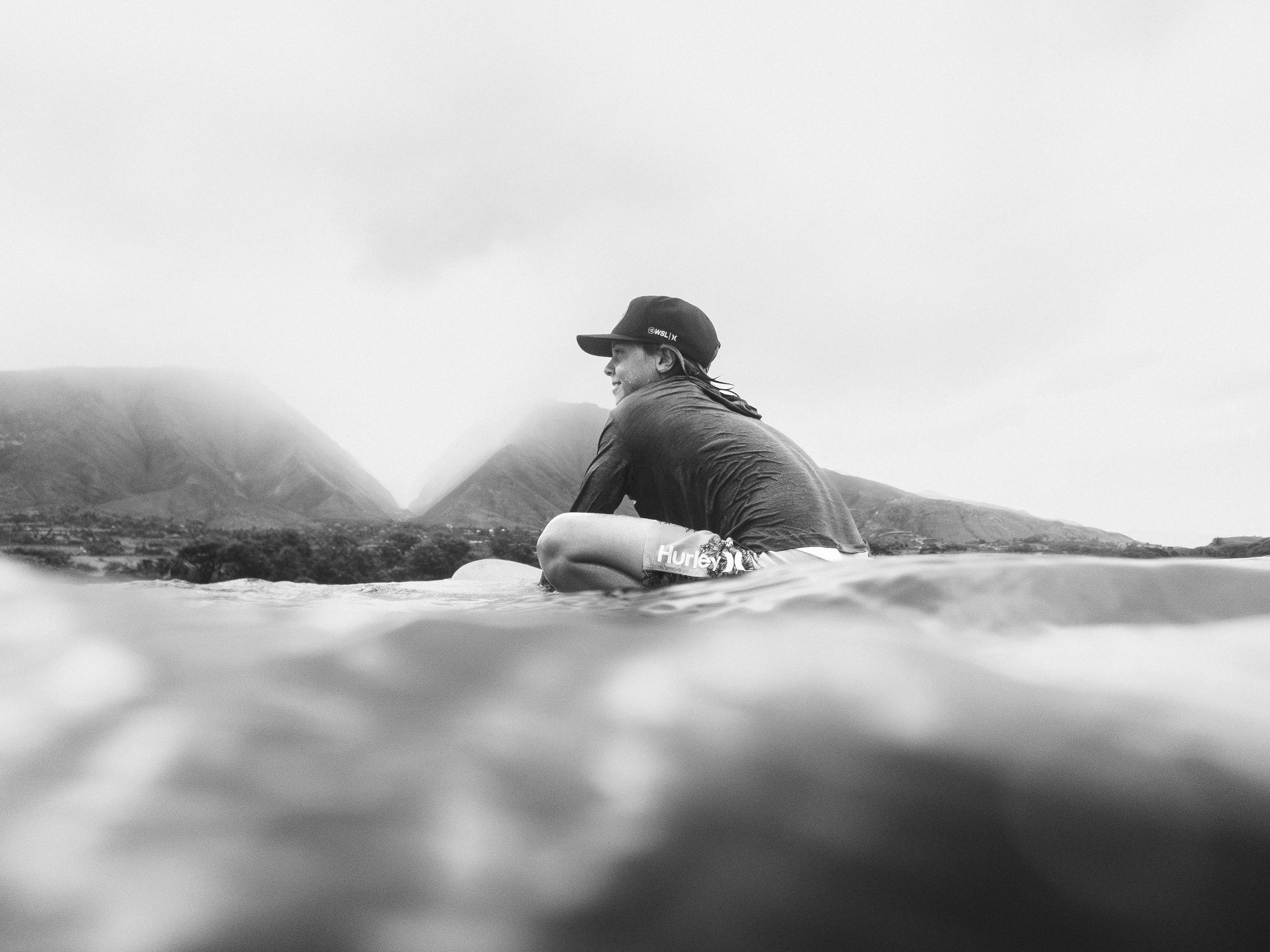Black and white photograph of a smiling person in a baseball cap and casual clothing sitting outdoors with mountains in the background, taken from a low angle at water level.