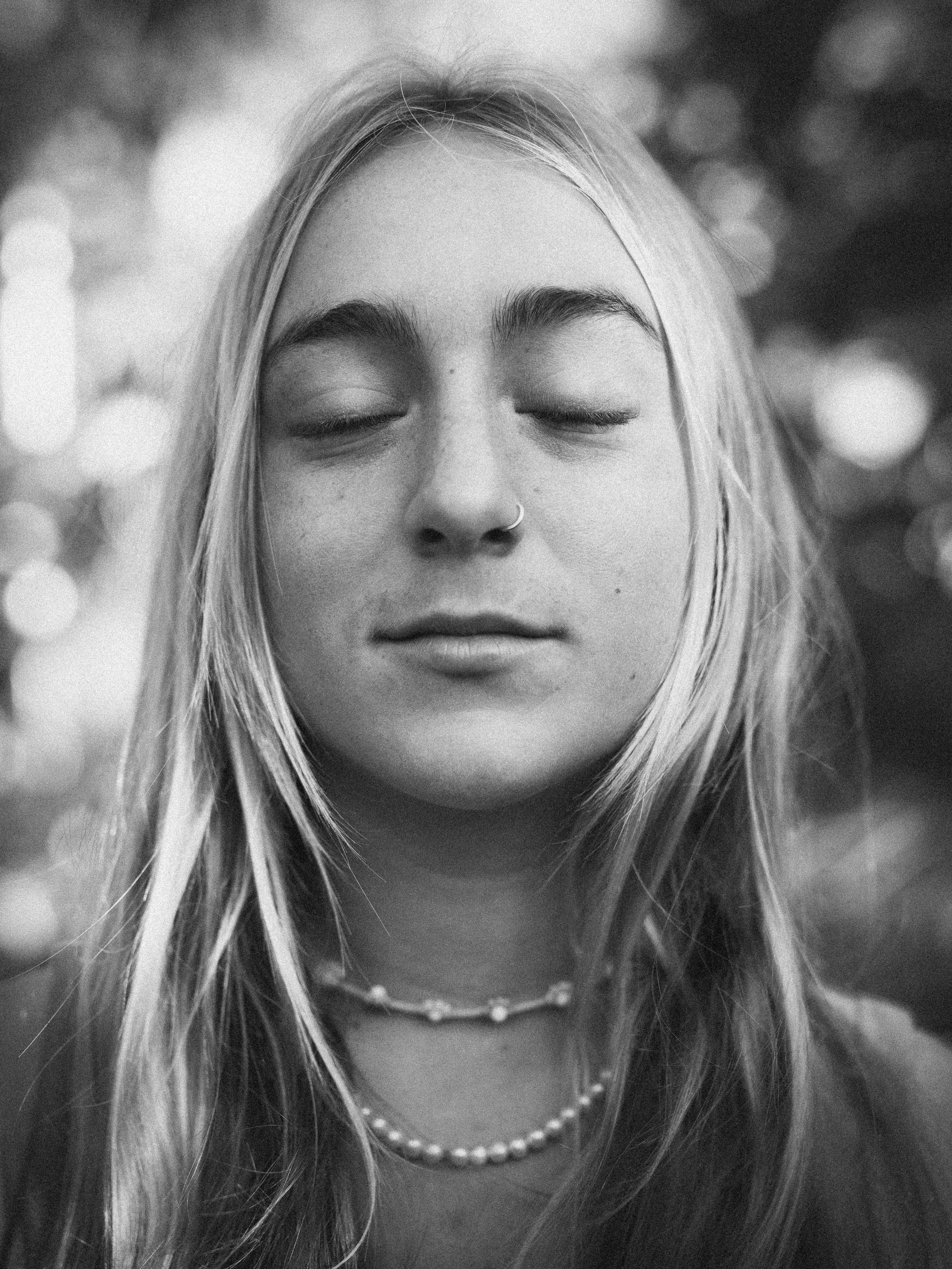 Close-up of a young woman with closed eyes, wearing a pearl necklace, a nose ring, and a delicate chain necklace, with blurred background.