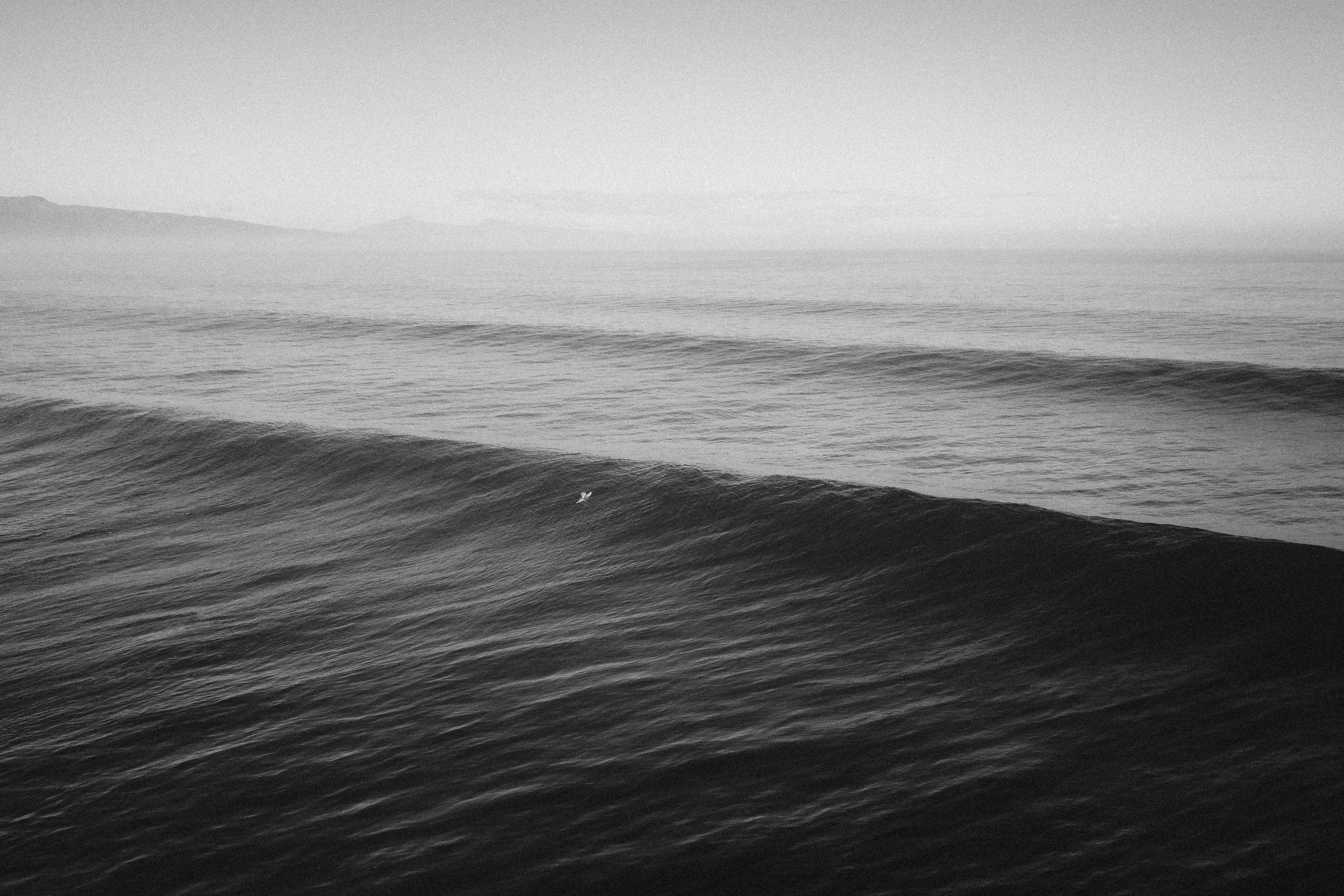 Black and white photo of ocean waves with mountain in the background.