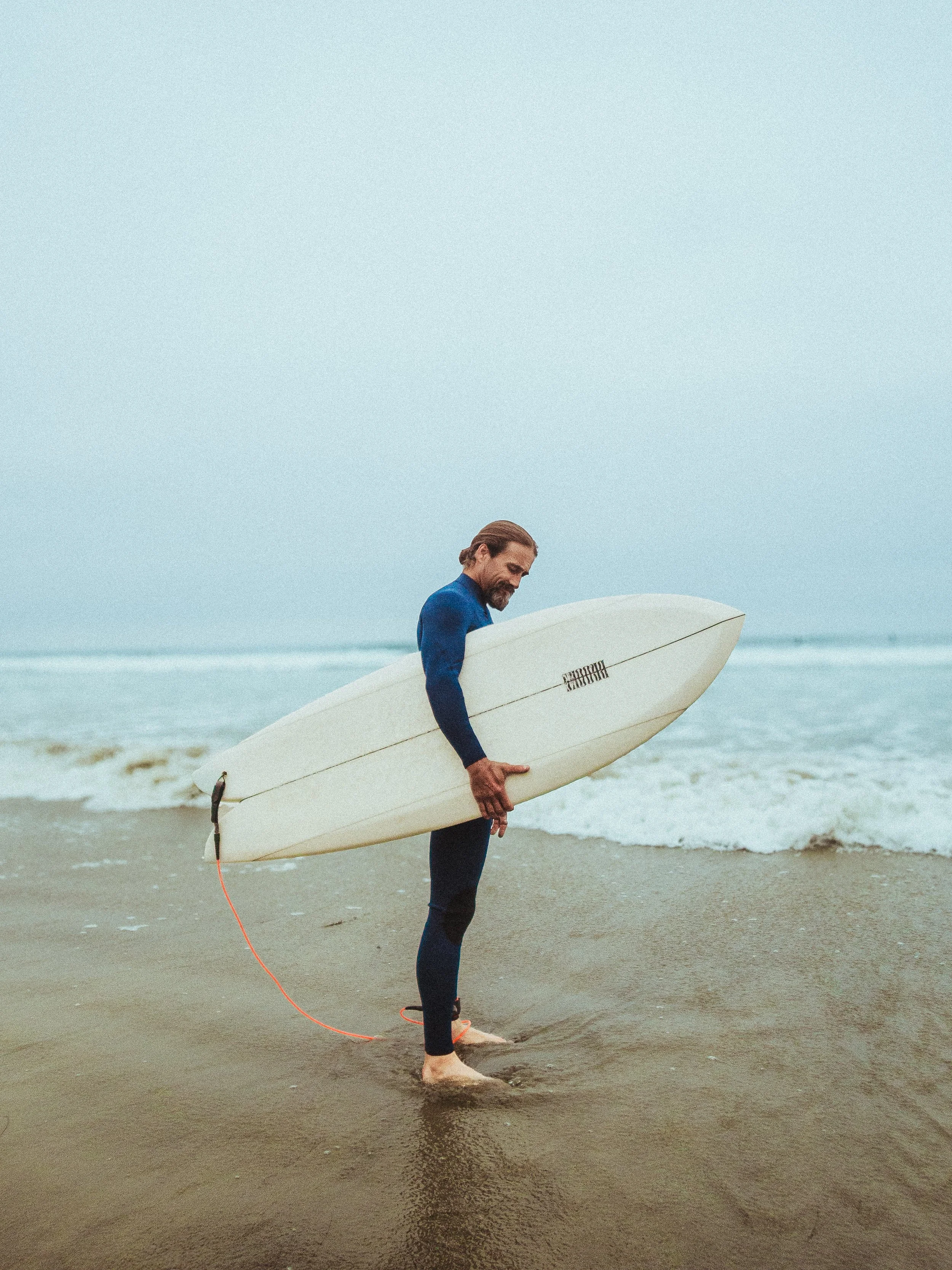 A man with long hair and a beard, wearing a blue wetsuit, standing barefoot on the beach holding a surfboard, looking down, with ocean waves in the background.