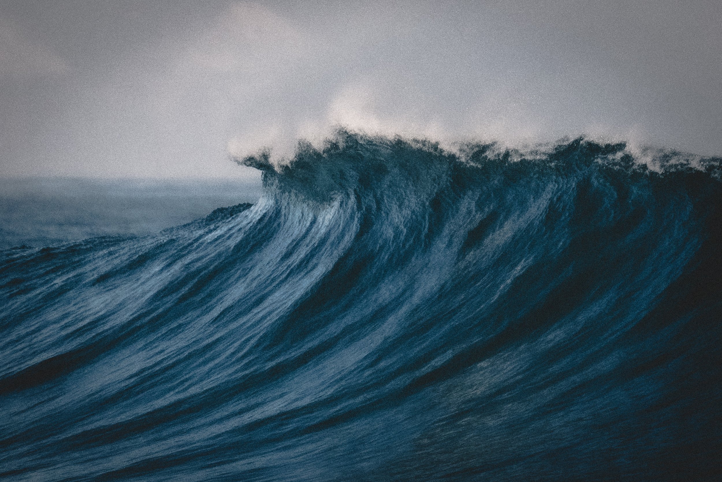 Large ocean wave with frothy white water cresting against a gray sky.