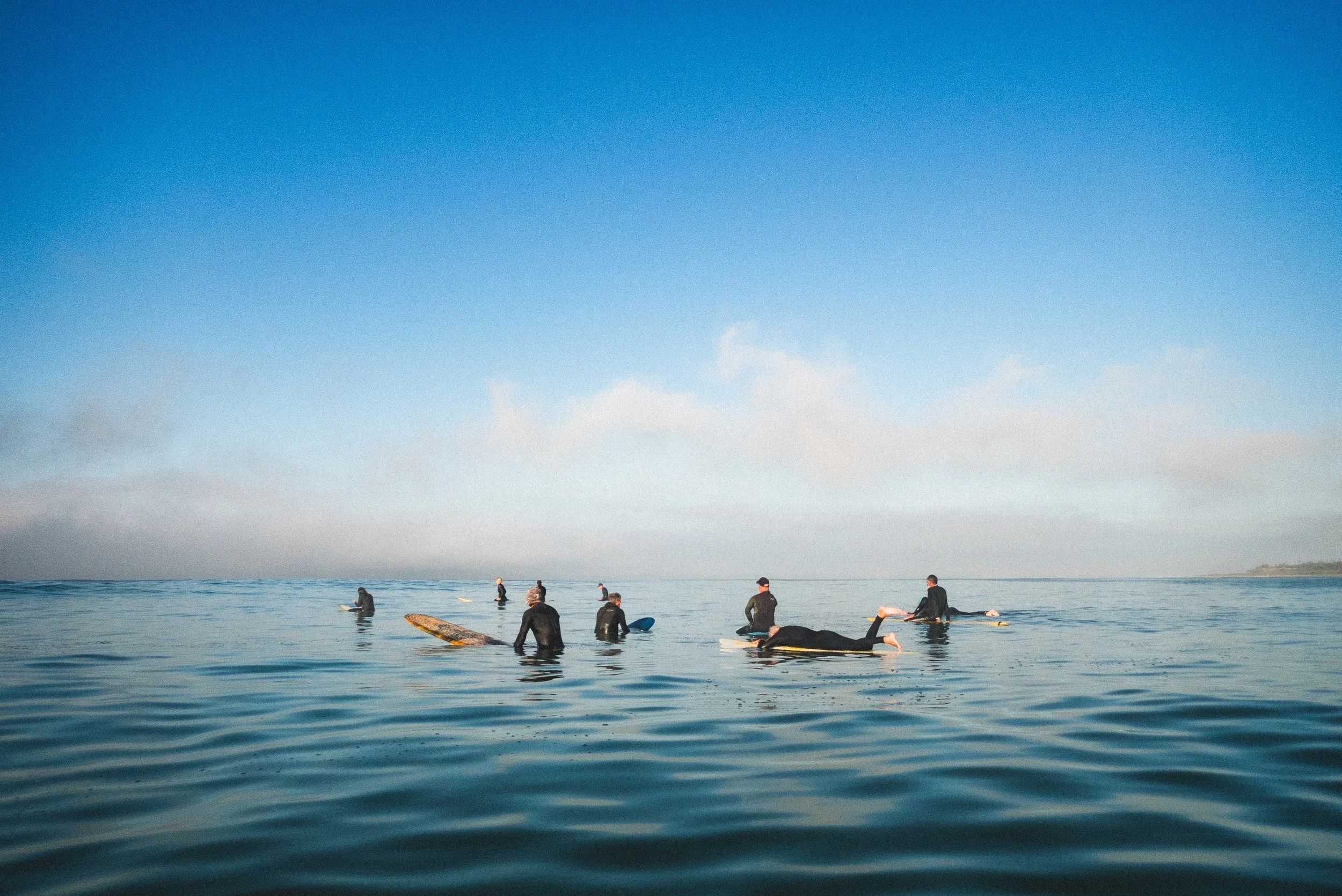 Several people are in the ocean, some sitting on surfboards and others standing or kneeling, under a mostly clear sky with a few clouds.