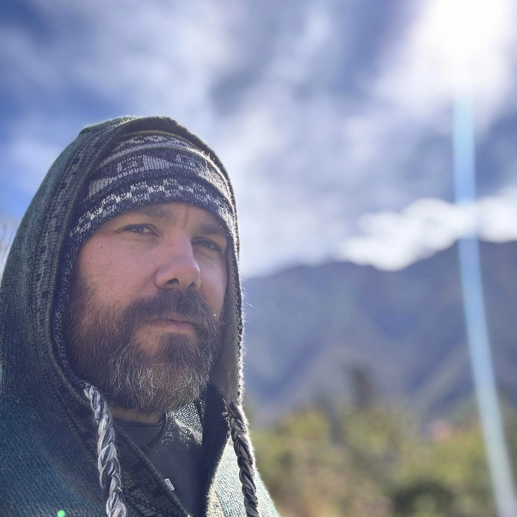 A bearded man wearing a winter hat with ear flaps and a hoodie outdoors on a sunny day with mountains and clouds in the background.