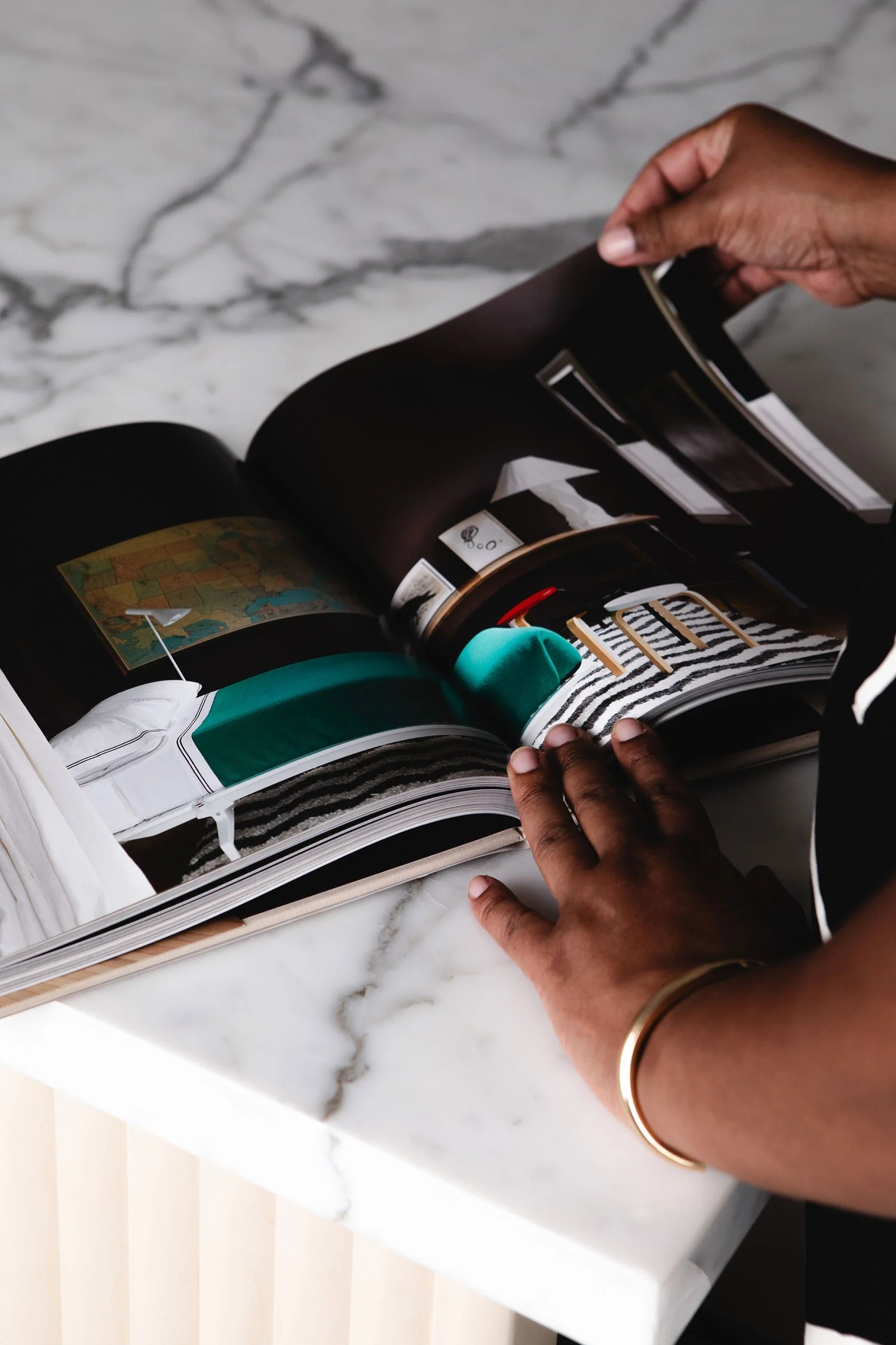 Person examining an open magazine with interior design images on a white marble table. The person has medium brown skin and is wearing a gold bracelet on their wrist.