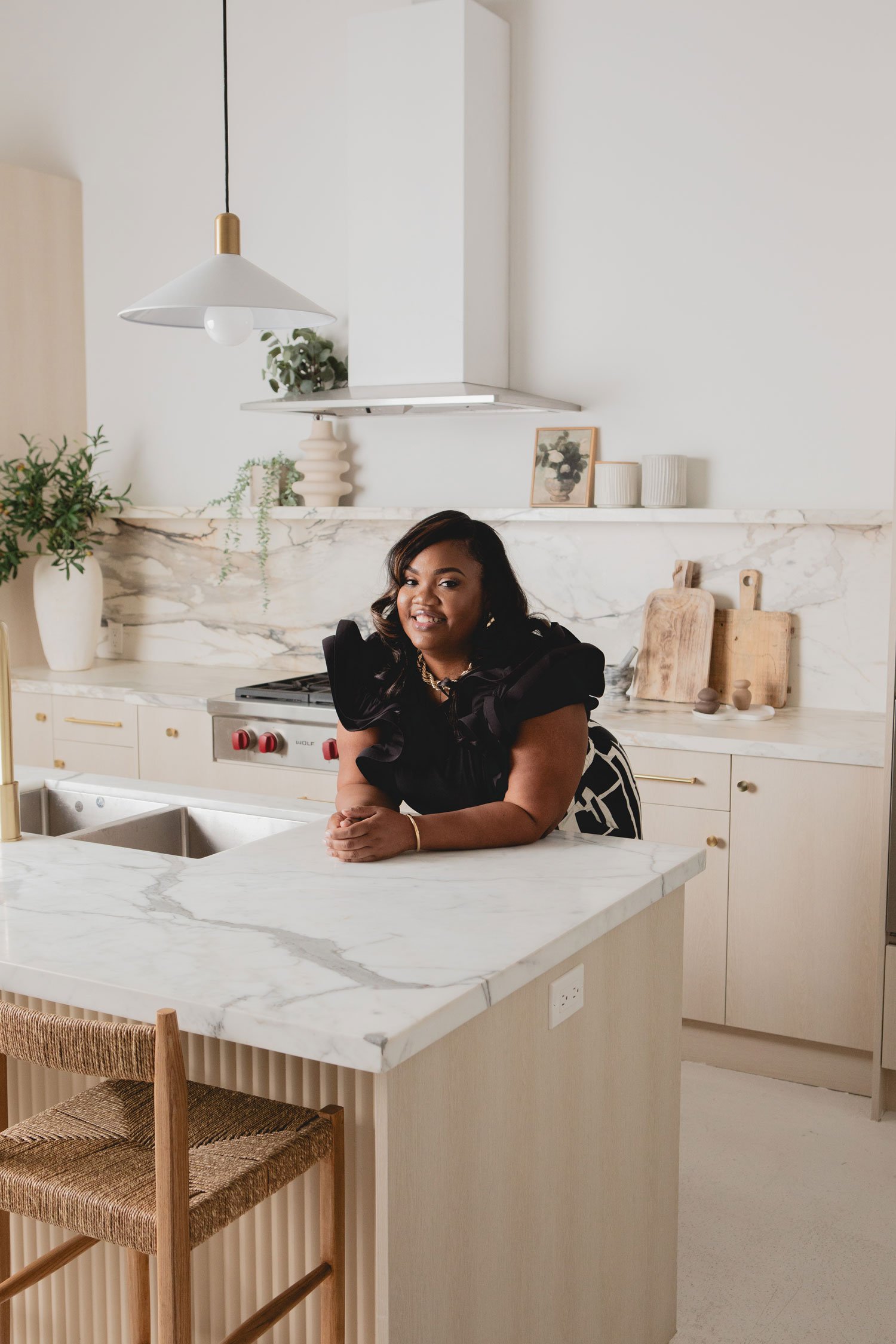 A woman with dark hair in a black ruffled top sitting at a kitchen counter made of white marble. The kitchen has white cabinets and a marble backsplash with minimalist decor, including framed artwork, potted plants, and cutting boards. There are wooden barstools with woven seats at the marble island.