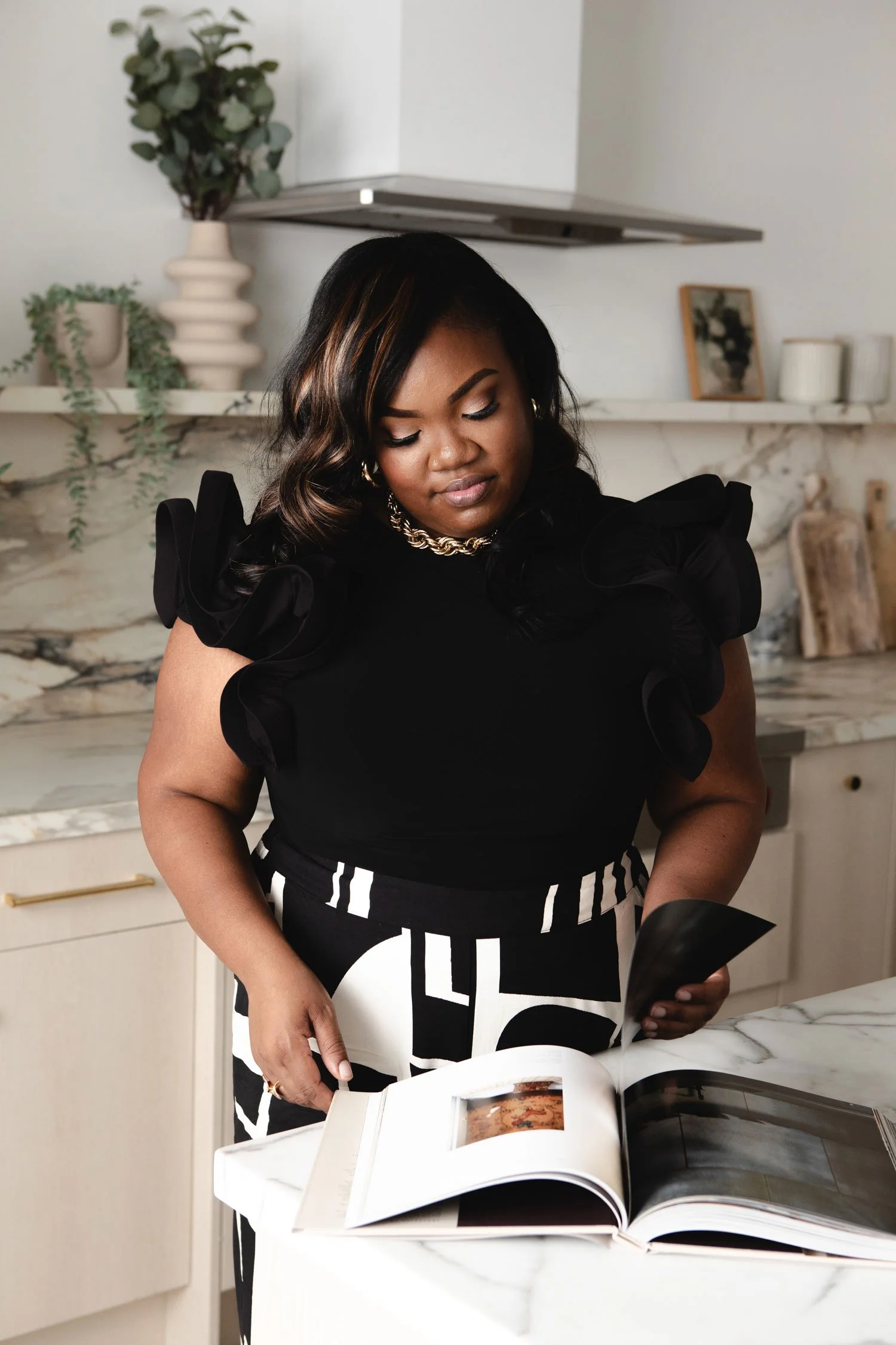 A woman with dark hair and a black top with ruffled sleeves looking down at a large open photo book on a white marble countertop in a modern kitchen with decorative vases and picture frames on the shelves behind her.