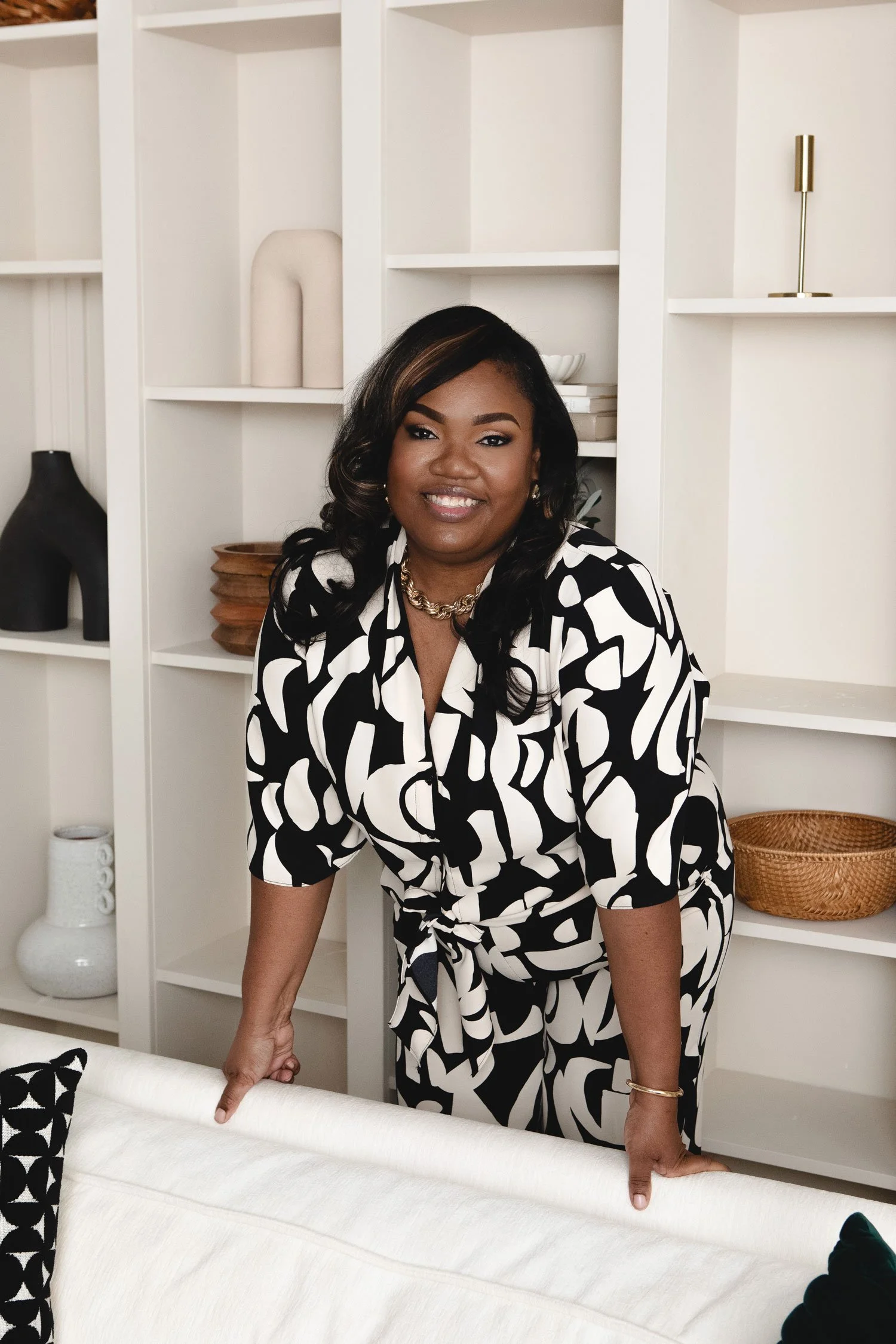 A smiling woman in a black and white patterned dress leans on a white sofa with her hands, in a room with white shelves decorated with bowls, vases, and sculptures.