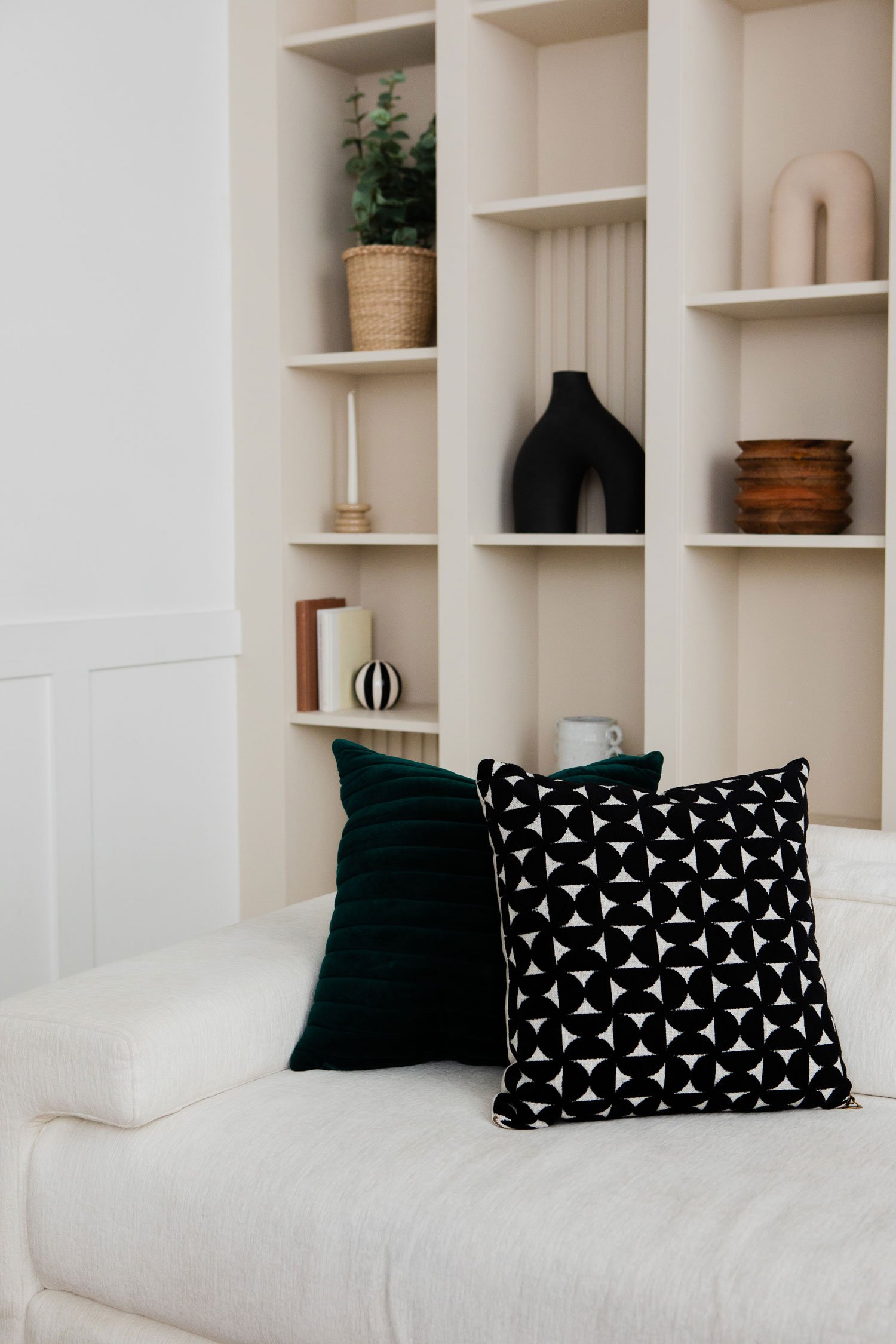 A white sofa with two black and white patterned pillows and a solid black pillow in front of a built-in white bookshelf with decorative items, including a potted plant, vases, bowls, and books.