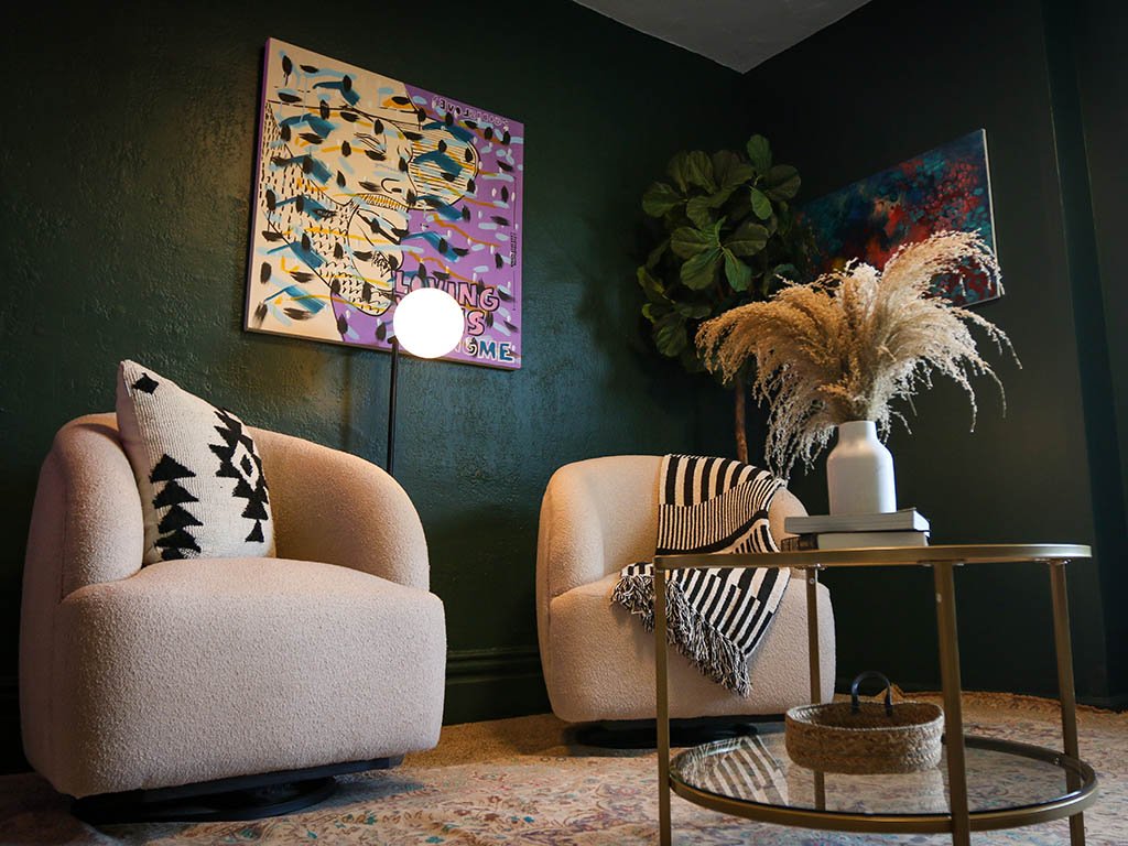 A cozy living room corner with two cream-colored armchairs, one with a black and white decorative pillow and the other with a striped throw blanket. Between the chairs is a gold coffee table with a large white vase filled with pampas grass and a few books. Behind the chairs, there is a dark green wall with colorful abstract artwork and a potted plant. There is also a floor lamp with a round white shade.