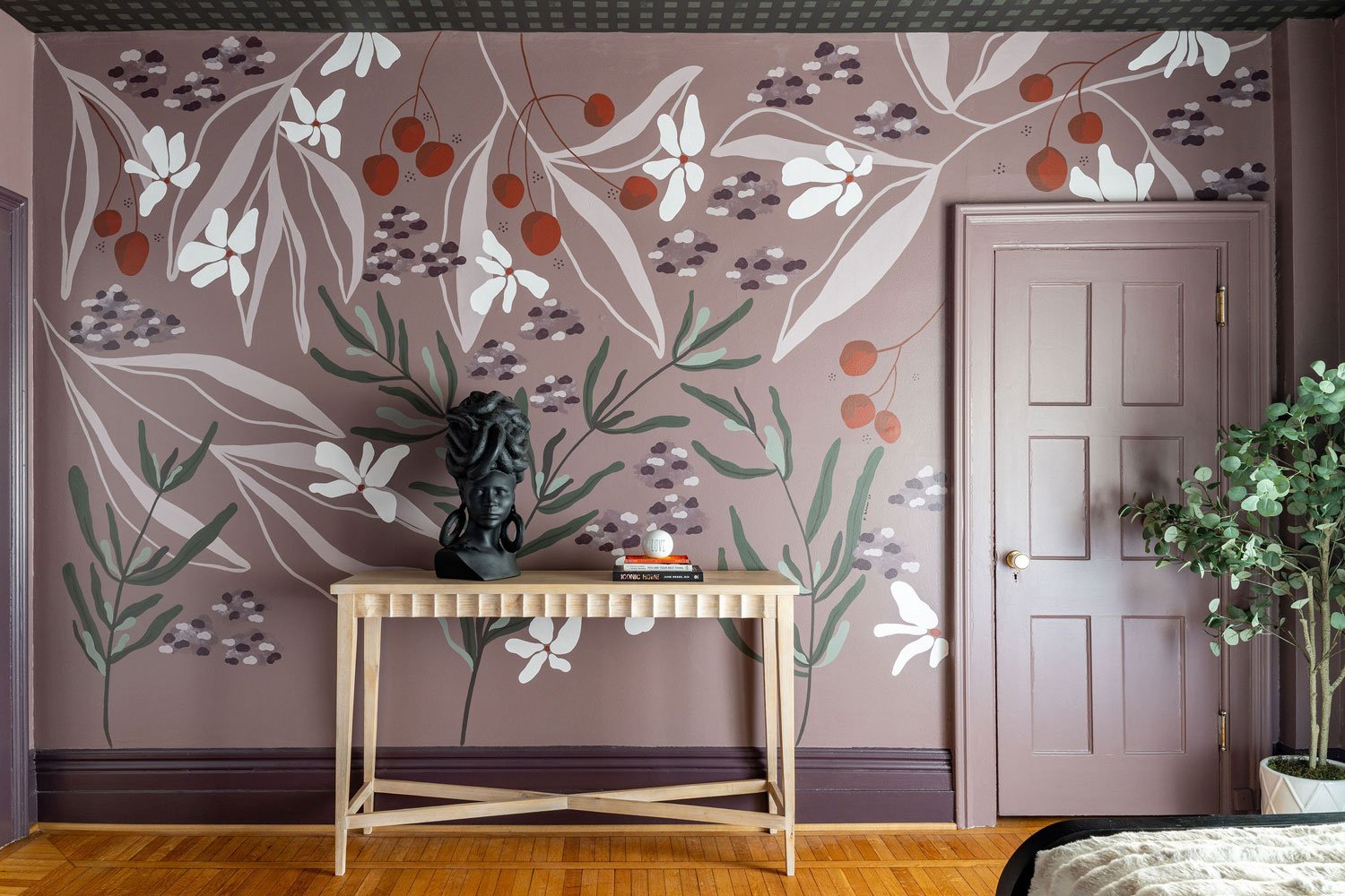 Interior room with a wall decorated with a floral mural featuring white flowers and red berries on grayish purple background. A wooden console table holds a black bust sculpture with dreadlocks and earrings, and a stack of books. There is a closed wooden door to the right and a potted plant with green leaves on the far right.