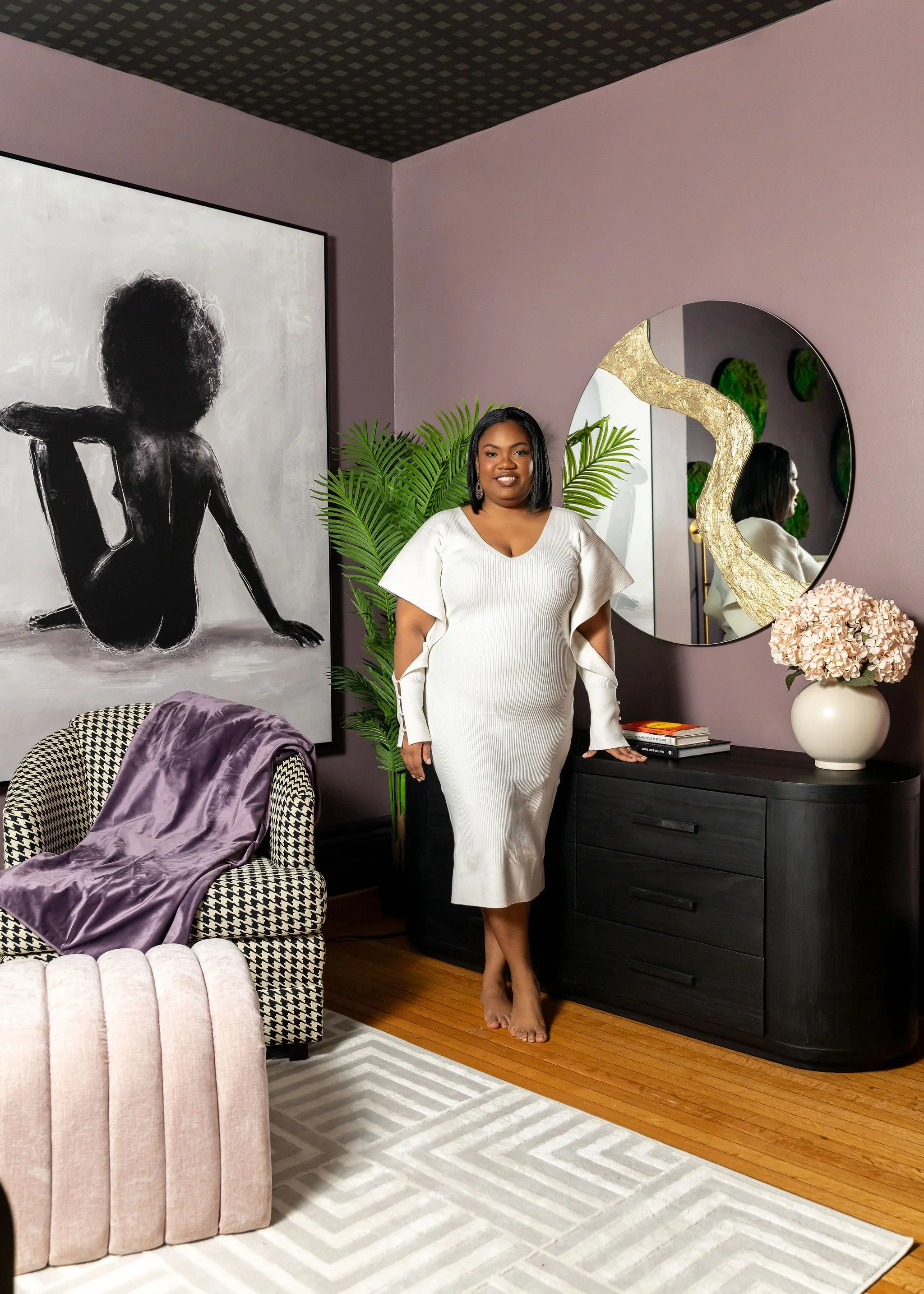 A woman in a white dress standing barefoot in a living room, beside a black sideboard with a large round mirror, pink flowers in a white vase, green plants, and artwork on a purple wall.