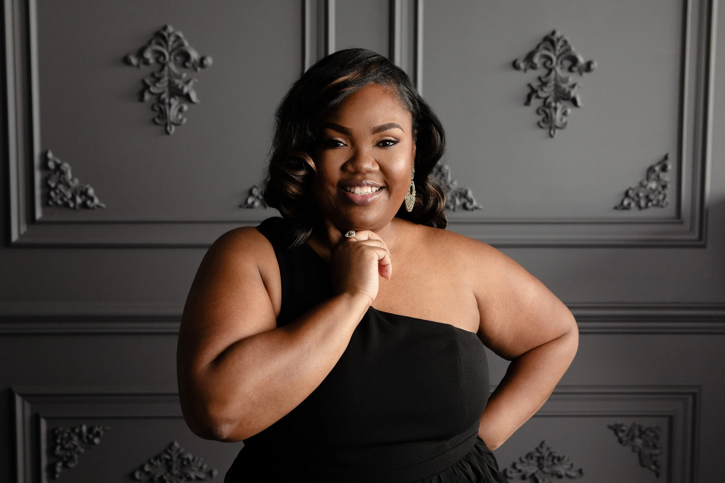 A woman with dark hair styled in loose waves, wearing a black dress and large, sparkling earrings, posing with her hand under her chin, in front of a dark gray wall with ornate black wall decor.