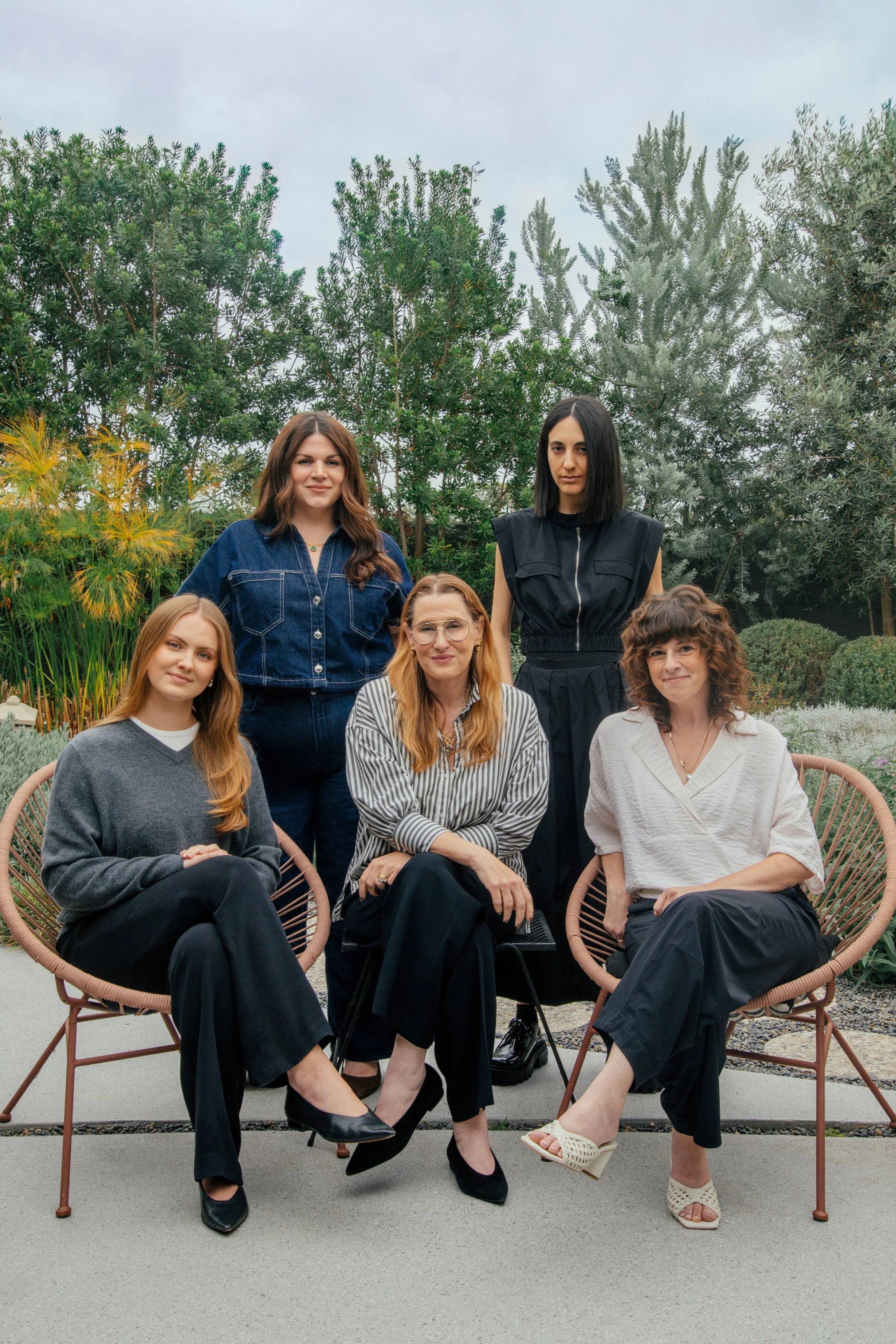 A group of six women sitting and standing outdoors among greenery, including trees and bushes. They are dressed in casual to semi-formal clothing and are sitting on pink, modern, circular chairs. The sky is cloudy.