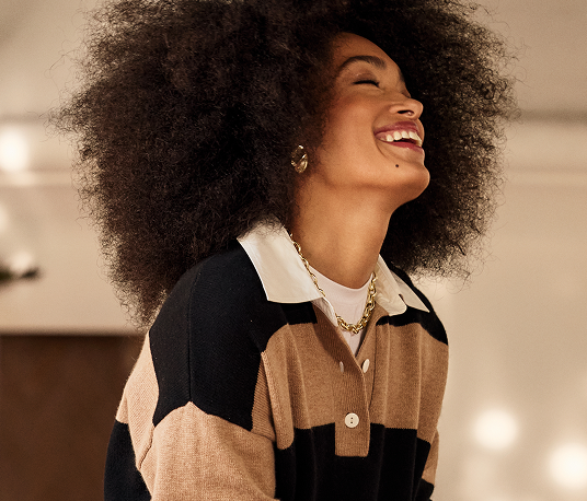 A woman with curly hair smiling and laughing, wearing a color-blocked sweater and gold jewelry.