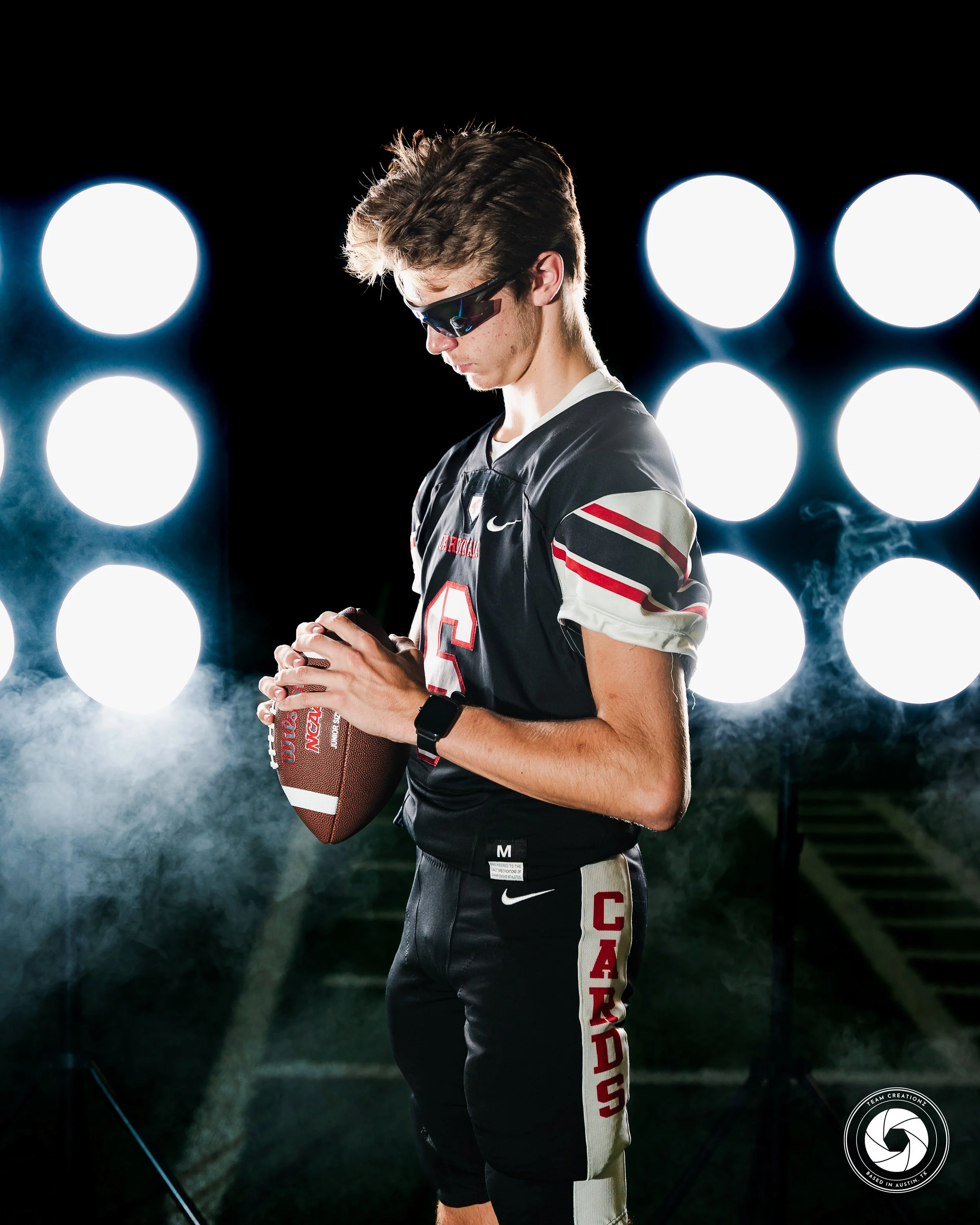 Football player in uniform holding a football, wearing sunglasses, with lights in the background.