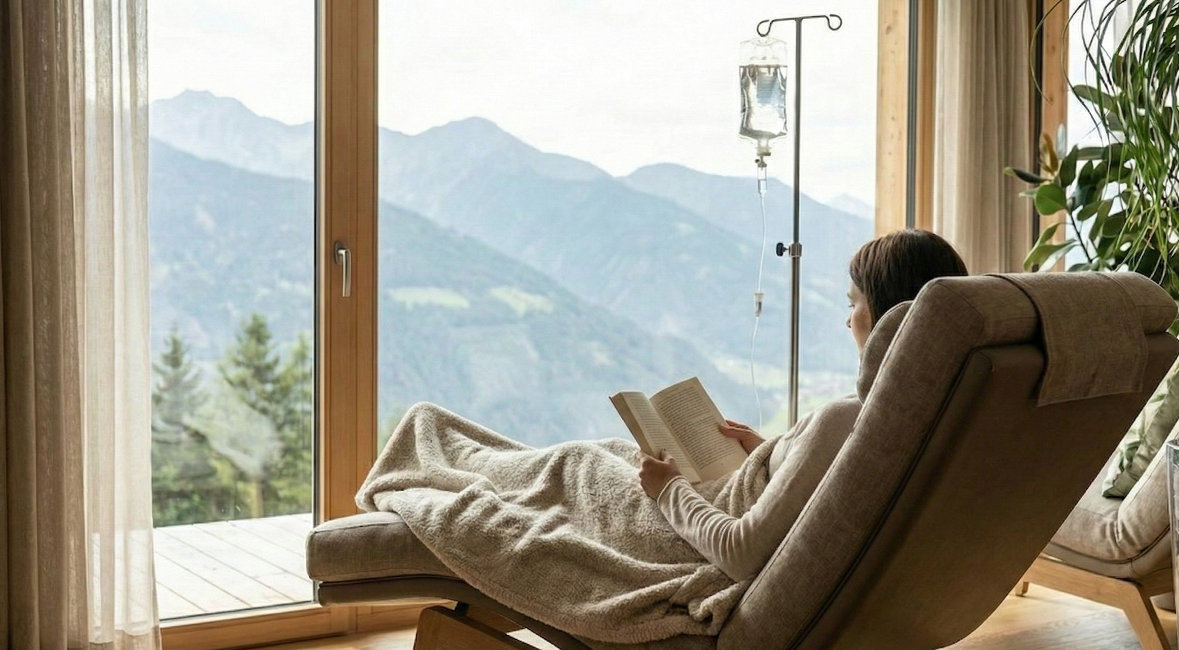 A woman sitting in a lounge chair, wrapped in a blanket, reading a book indoors next to a large window with mountains in the background, an IV drip stand nearby.
