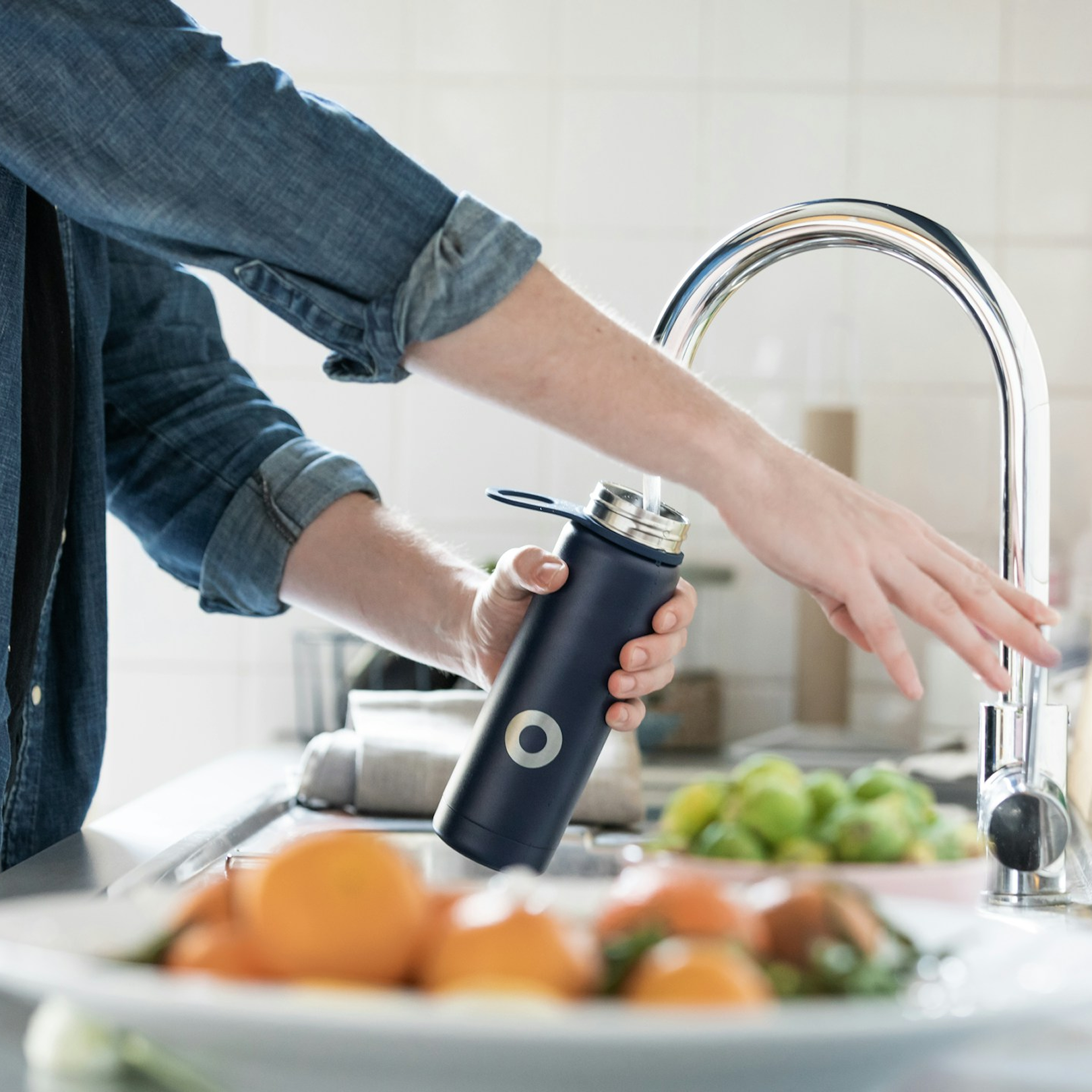 Person filling a black reusable water bottle at a kitchen sink with a modern chrome faucet, surrounded by fruits. They represent a Quench IV Drip patient benefiting from restored fluid balance, muscle contraction, acid-base balance, and hydration.