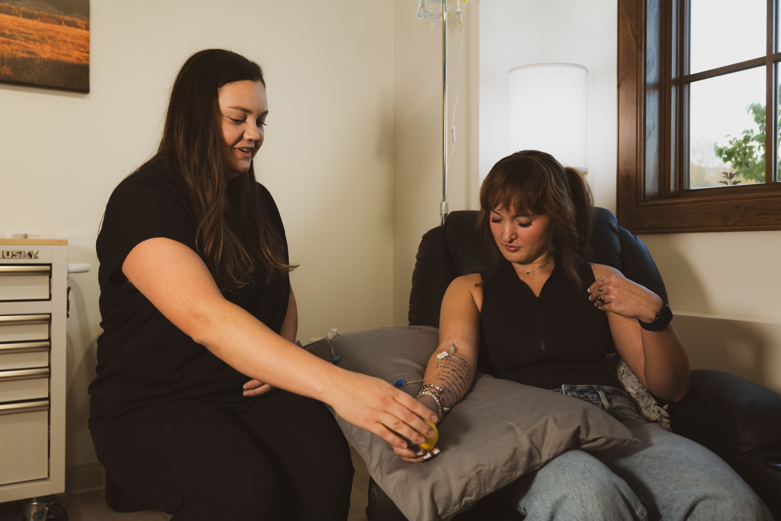 A woman receiving an IV therapy drip in her arm from a licensed medical professional in a room with natural light.