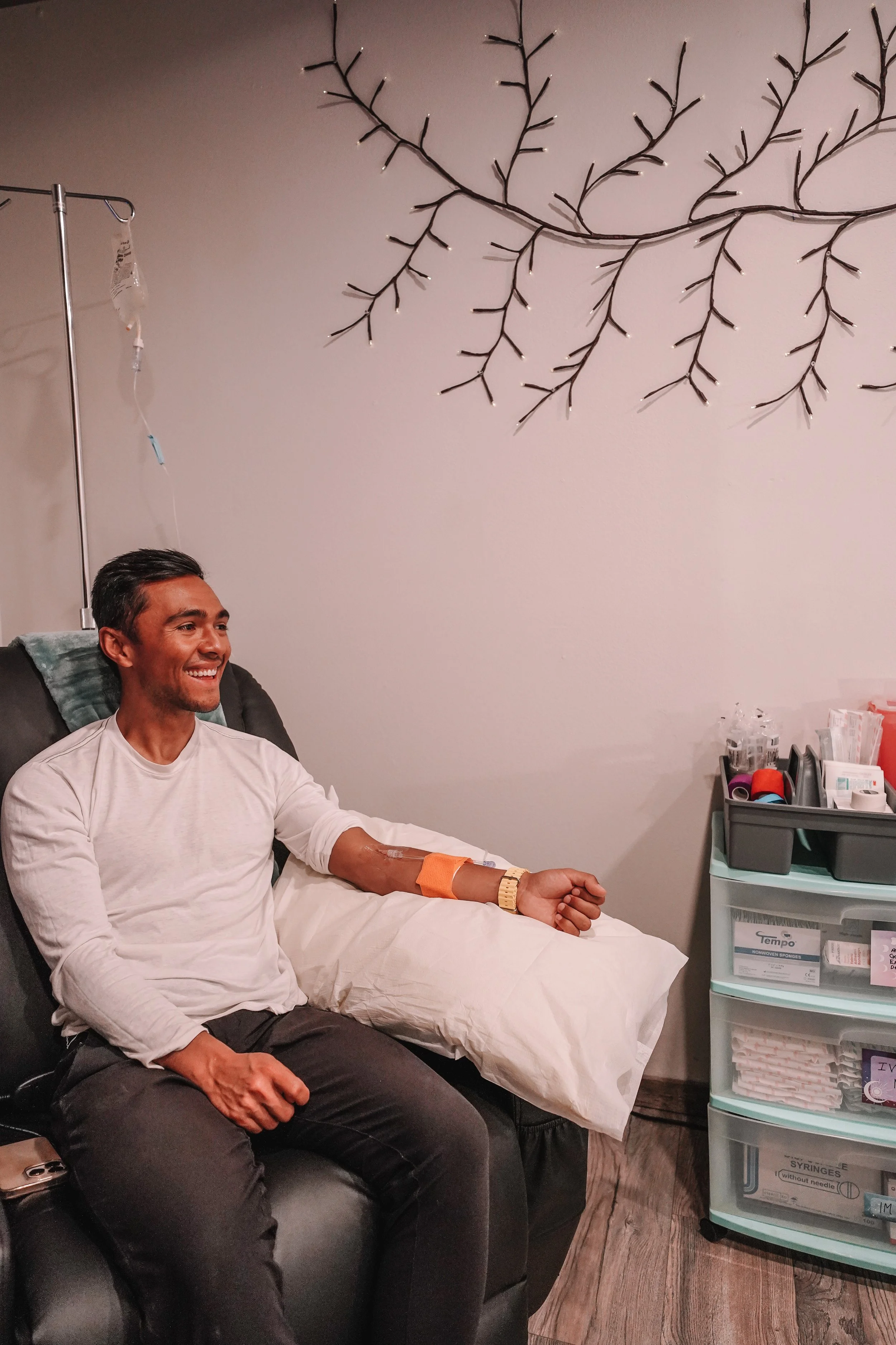 A man sitting in a medical chair after donating blood, smiling. His right arm has a bandage on it with a needle inserted for the blood donation. There is an IV fluid bag hanging on a stand on the left side, and medical supplies on a plastic drawer on the right.