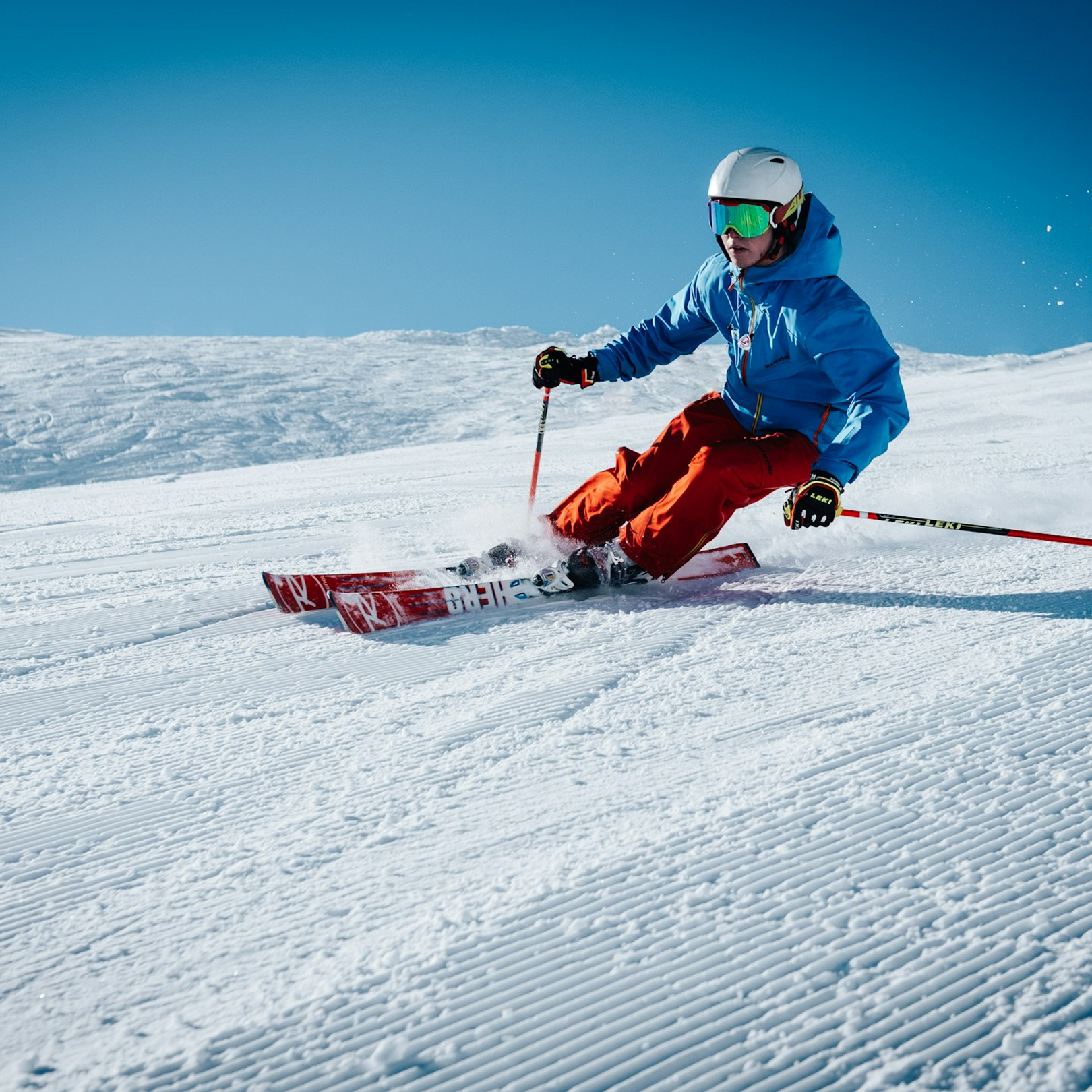 A skier in a blue jacket, orange pants, wearing a white helmet and goggles descends on a snowy slope. They represent a patient who receives the Athletic & Performance Recovery Iv drip which supports performance and recovery.