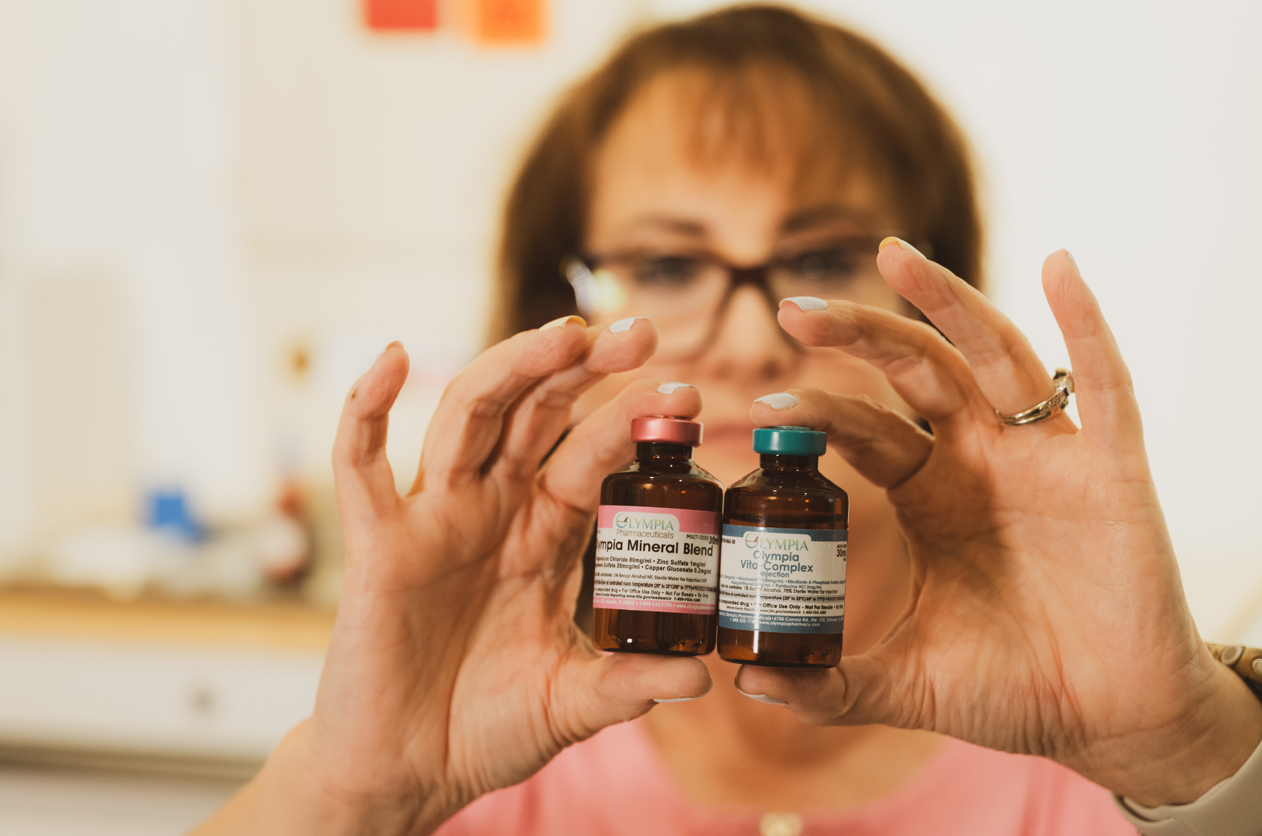 Deb, a licensed medical professional, holding two IM Injection bottles labeled 'Olympia Mineral Blend' and 'Olympia Vitamin Complex' in front of her face, with her hands positioned to display the bottles clearly.
