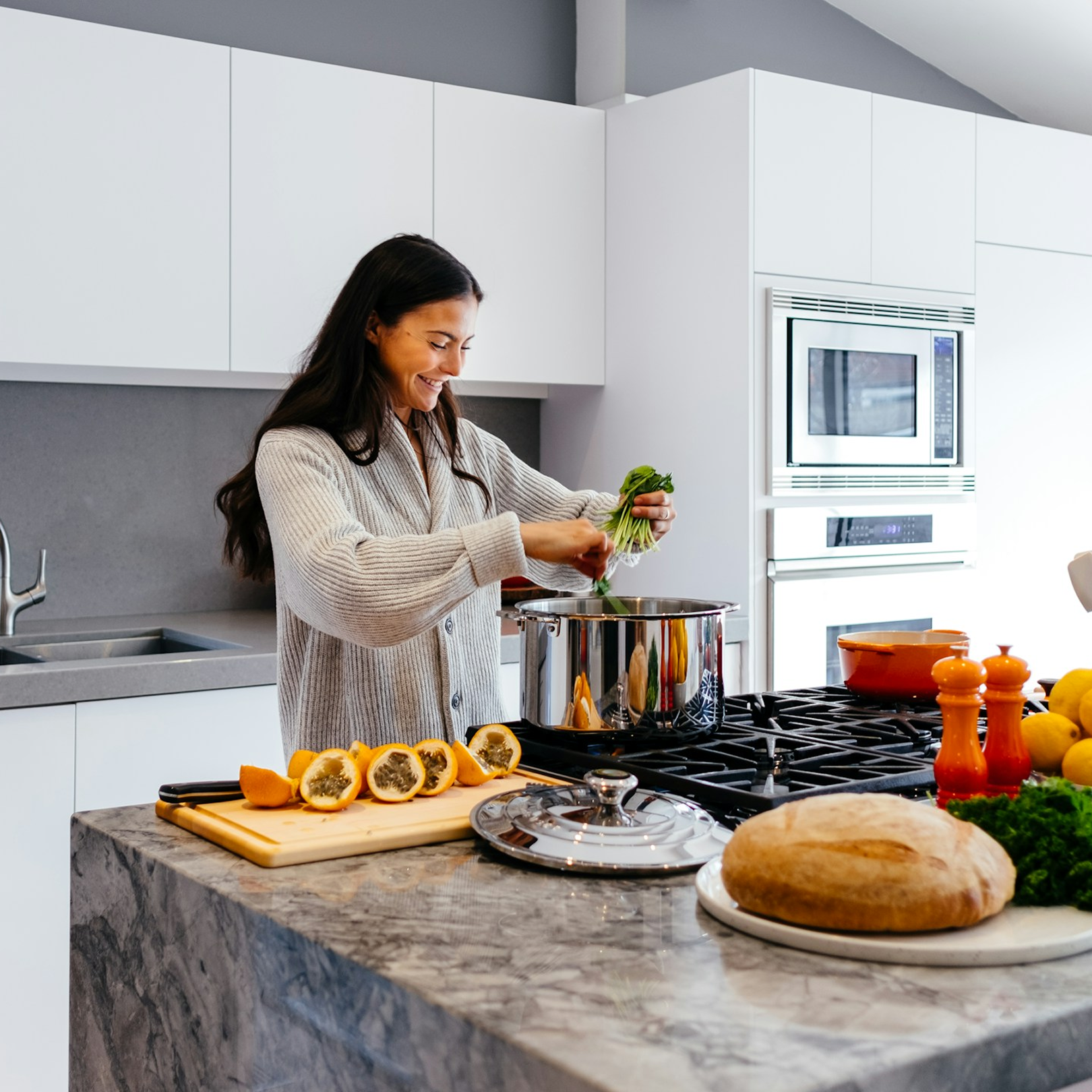 A woman cooking in a modern kitchen with a marble countertop, adding greens to a pot. She represents a patient who benefits from the High C IV drip which supports the immune system and benefits skin, bones, and cardiovascular health.