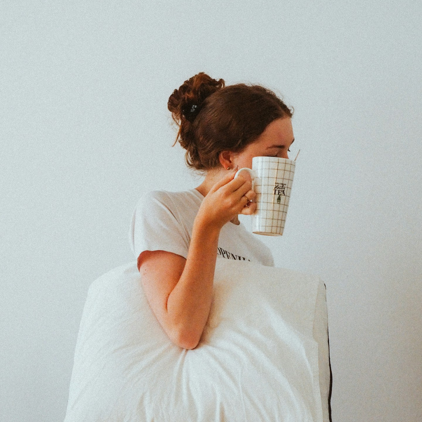 Woman sitting on a couch, drinking tea from a plaid mug, with a pillow on her lap, against a plain white wall. She represents a Hangover IV Drip patient benefiting from a blend of fluids, electrolytes, and vitamins to combat hangover systems.