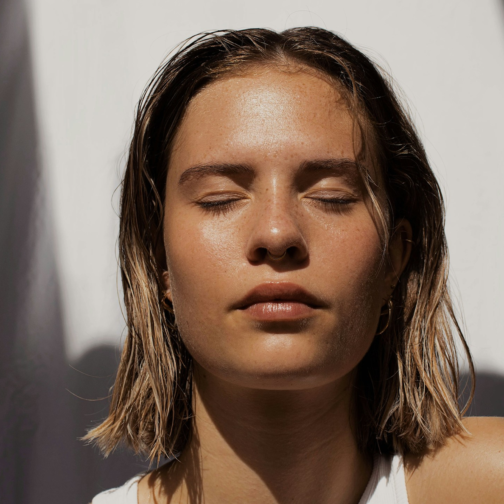 Close-up of a young woman with wet hair, closed eyes, and natural makeup, facing the sunlight with a serene expression. She represents a Radiance IV Drip patient benefiting from a youthful glow from improved complexion and collage production.