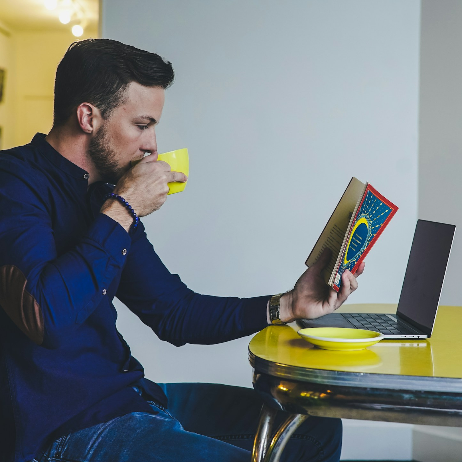 A man sitting at a yellow table, drinking from a yellow mug while reading a book, with a laptop open in front of him. He represents a patient  who benefits from the Cognitive IV drip which supports cognitive decline.