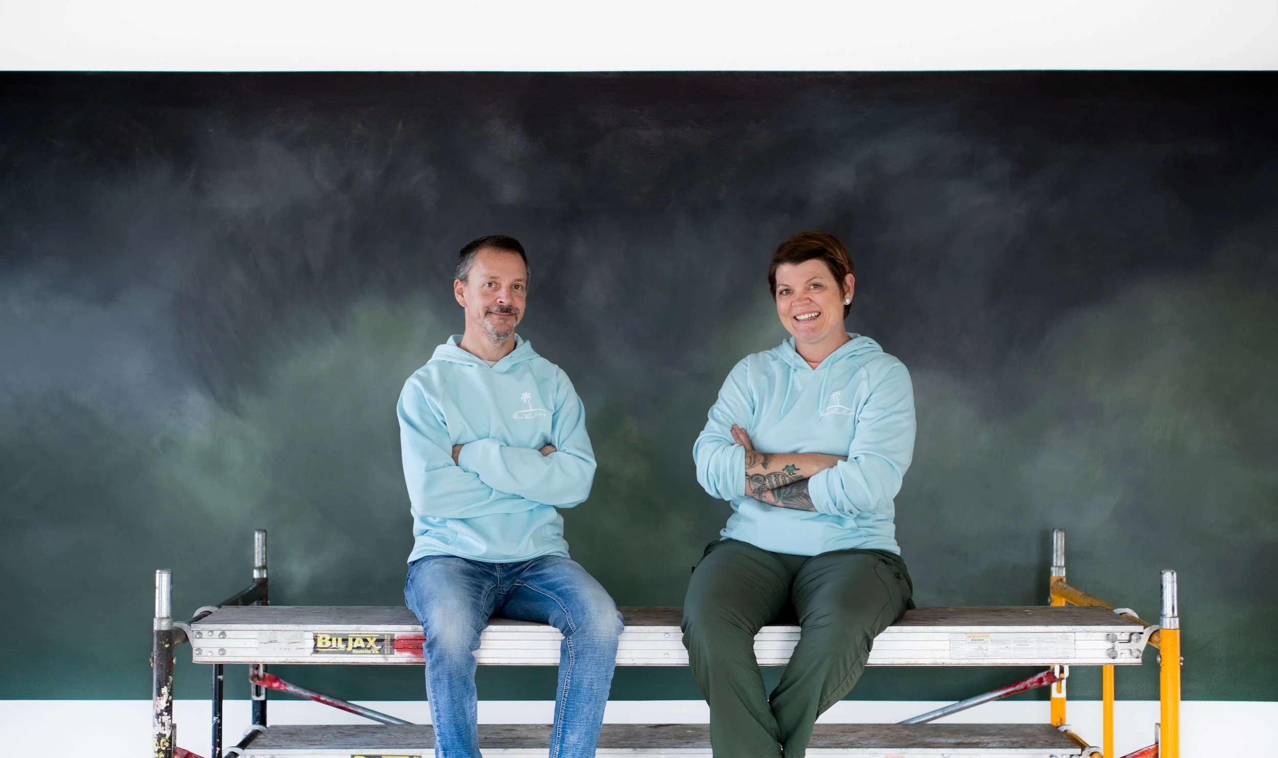 Two people sitting on a scaffold in front of a dark green wall, both wearing light blue hoodies, smiling and looking at the camera.