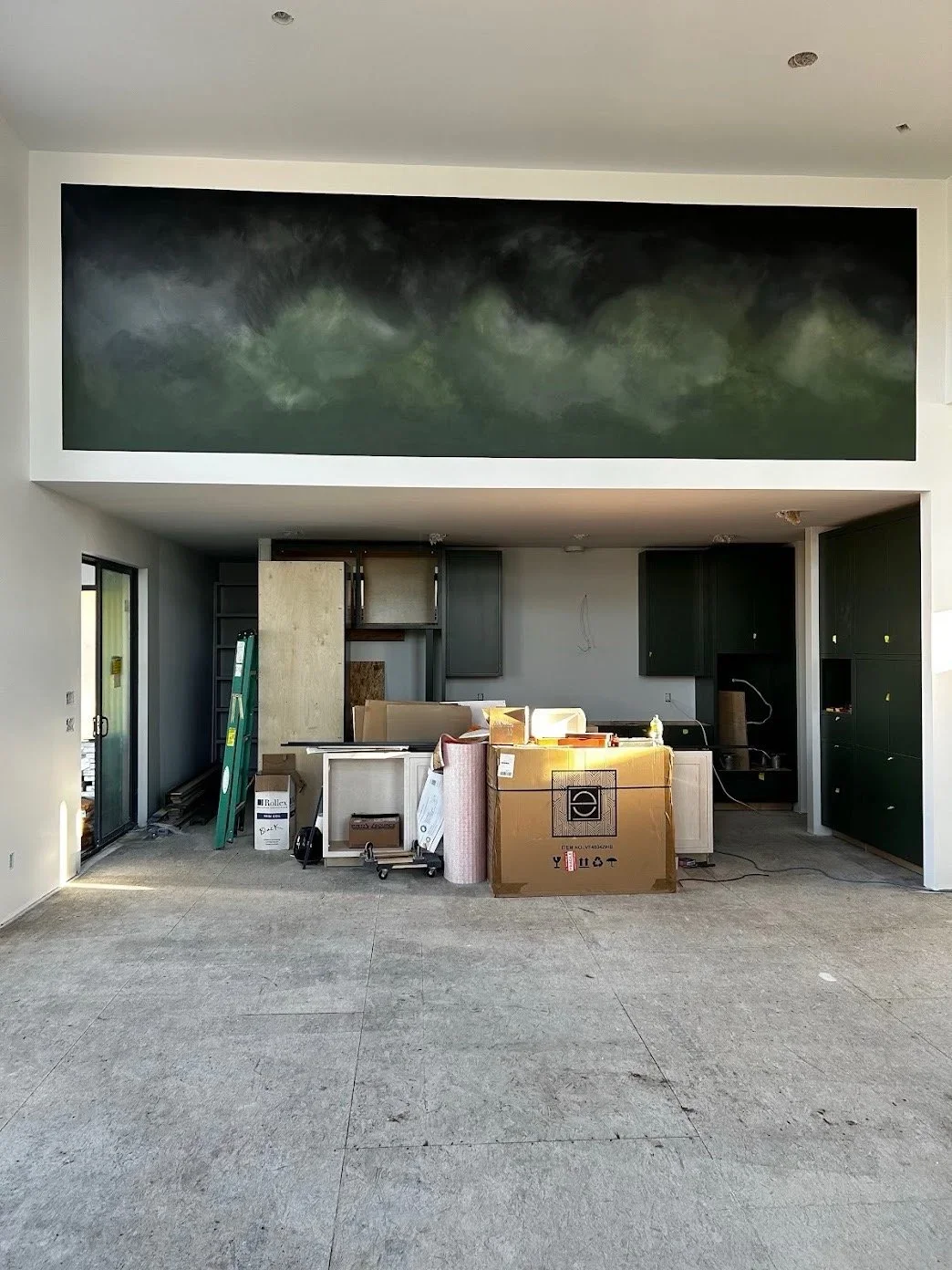 Interior view of a room under construction with cabinets, boxes, and construction materials, and a large painting or mural of a stormy sky or clouds on the upper wall.