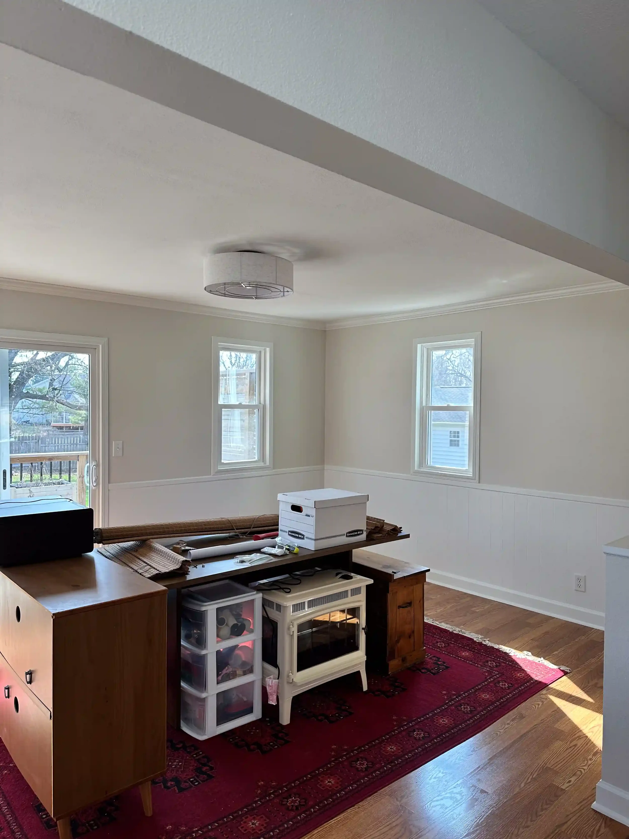 Interior of a living room with a red patterned rug, white walls, three windows, a ceiling light fixture, and a wooden cabinet with storage bins on the left side. Items are placed on the cabinet and the floor, indicating a space in the process of organizing or cleaning.