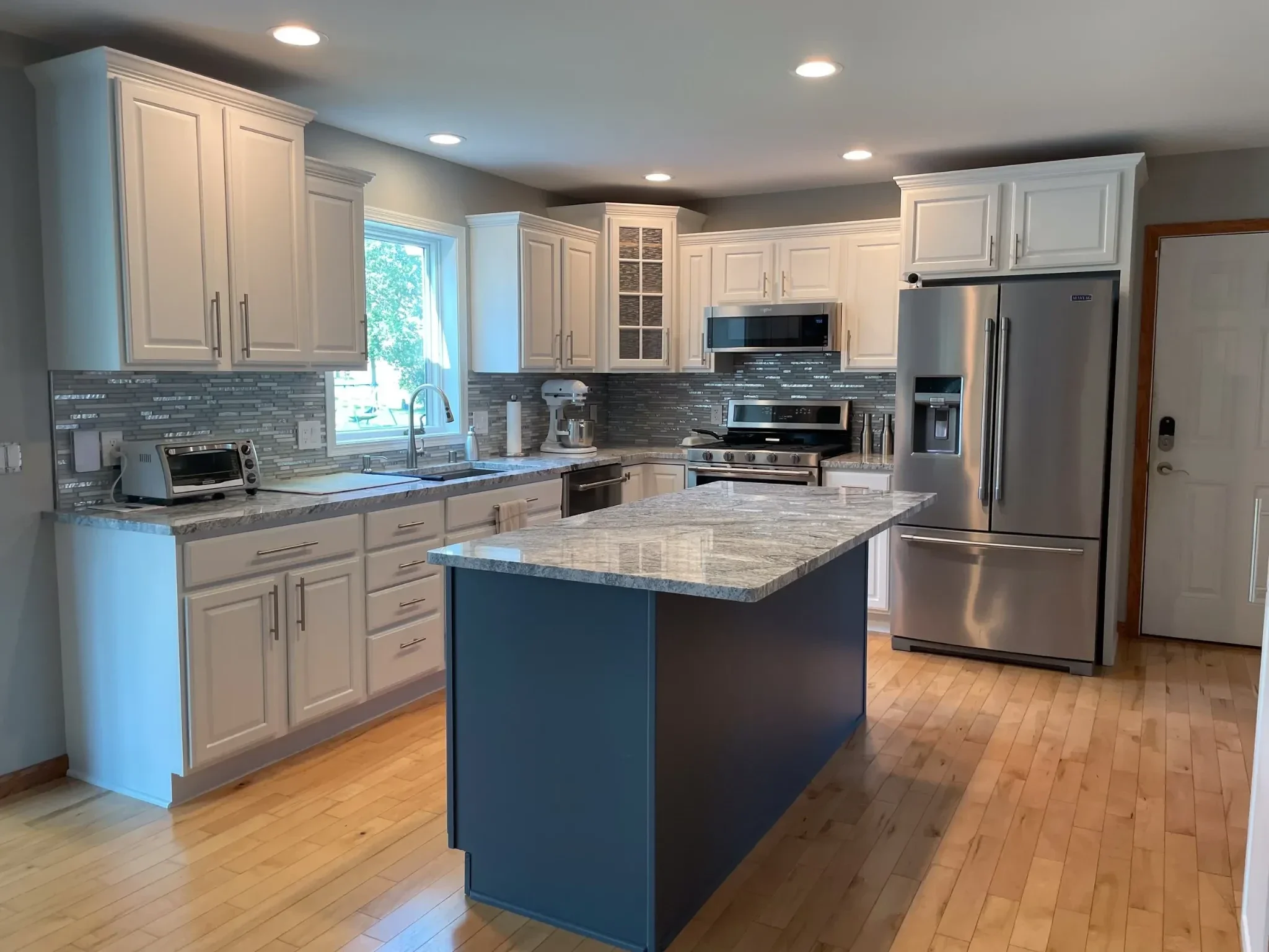 A modern kitchen with white cabinets, a granite countertop, stainless steel appliances, a kitchen island, a window with sunlight, hardwood flooring, and recessed ceiling lights.