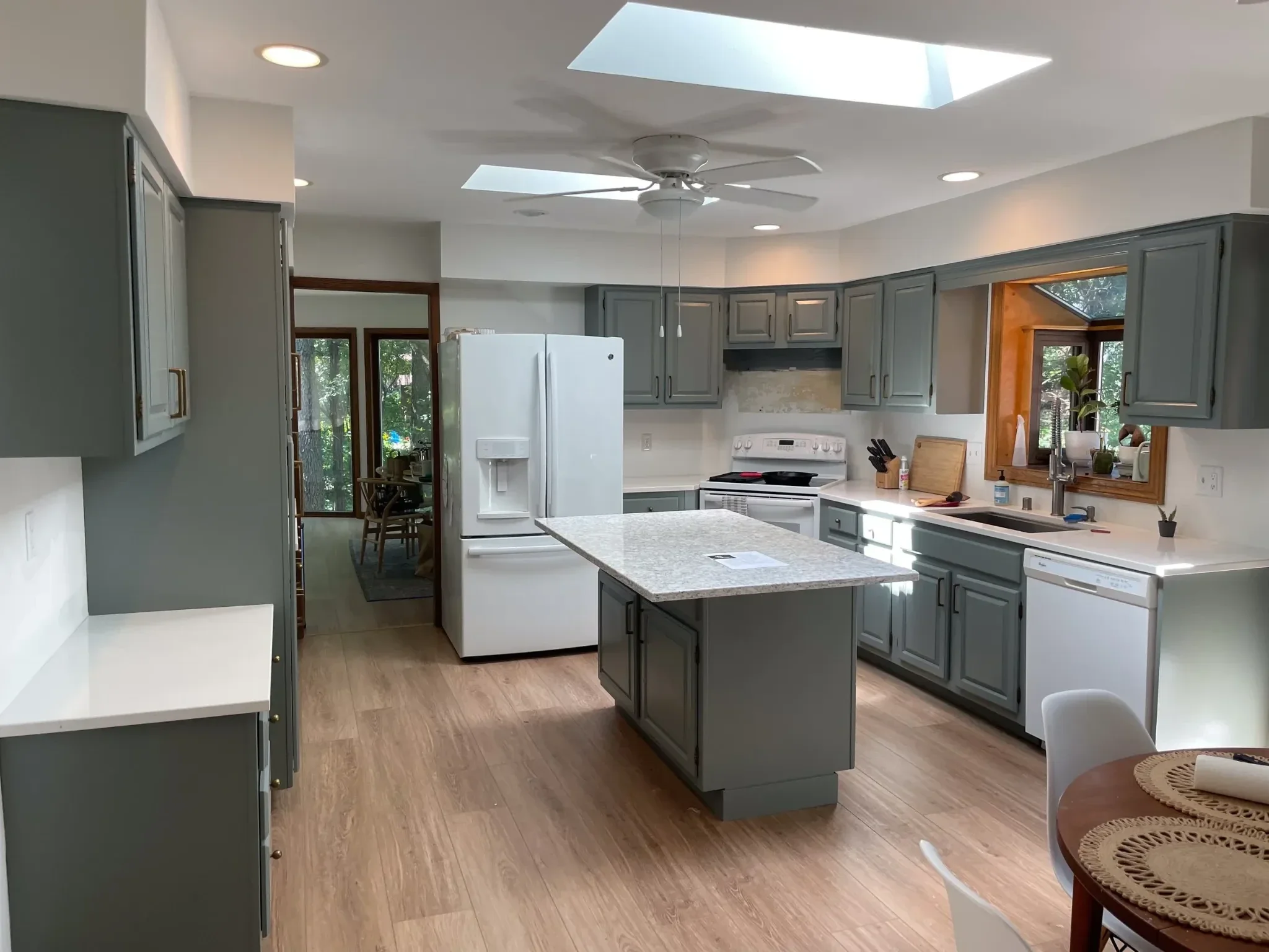 A kitchen with gray cabinets, a white refrigerator, a white stove, a kitchen island with a granite countertop, a ceiling fan, skylights, and a wooden dining table with chairs near a window.