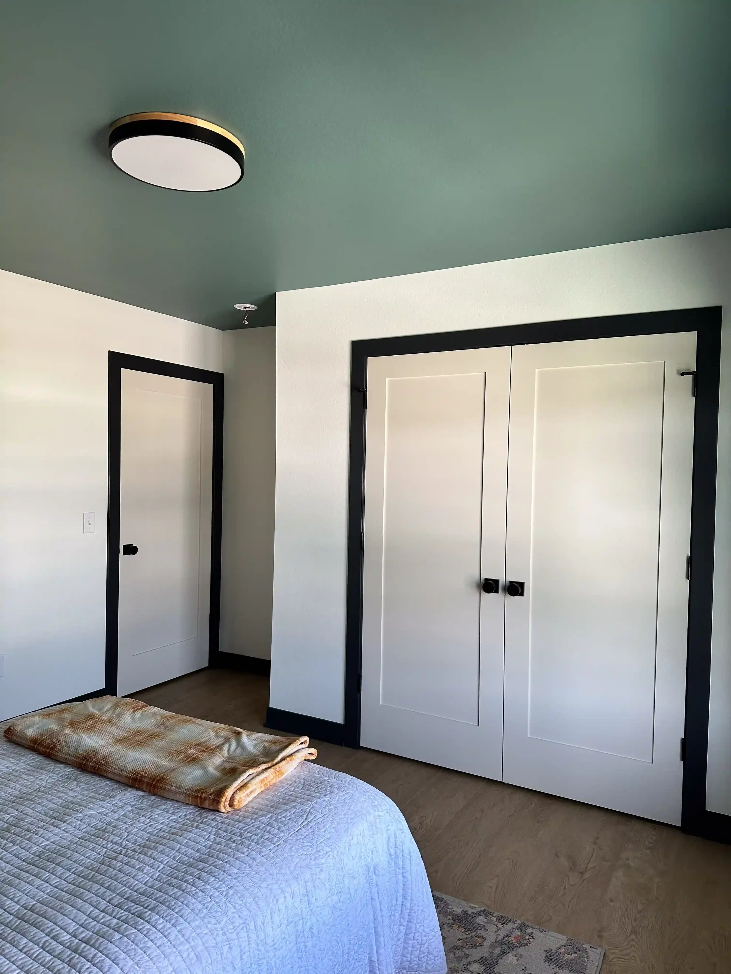 A bedroom with a white bedspread, folded beige and white blankets at the foot, a white wall, a black-trimmed closet with double doors, a white door with a black knob, and a wood floor with a small patterned rug.