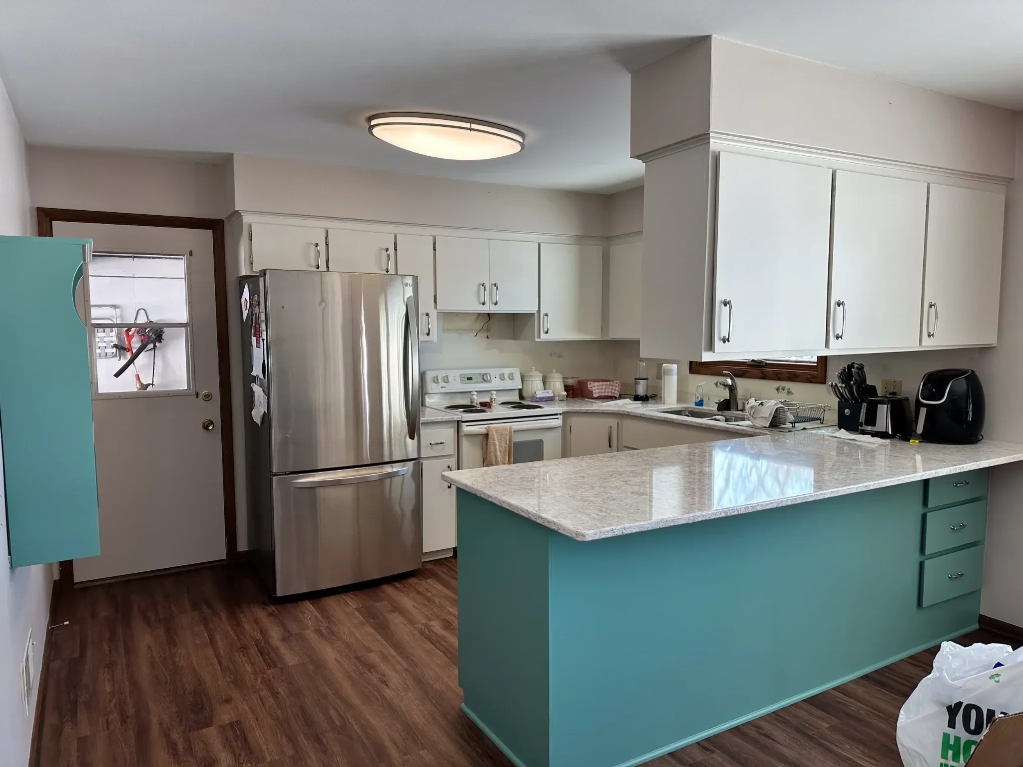 Kitchen with white cabinets, a stainless steel refrigerator, and a white stove. There is a white countertop island with teal cabinets underneath, and a kitchen sink. The ceiling has a round flush light fixture. The floor is dark wood.