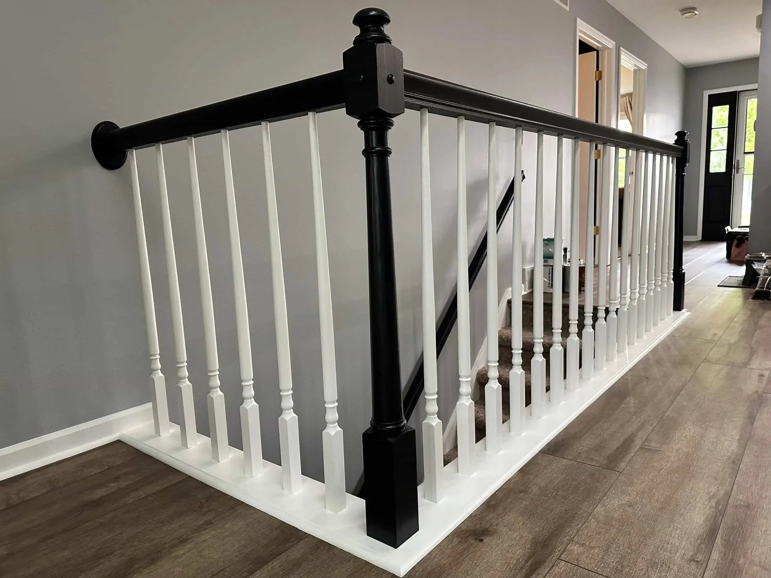 Interior view of a staircase with a black handrail, white balusters, and a black newel post, with a gray wall and a door in the background.