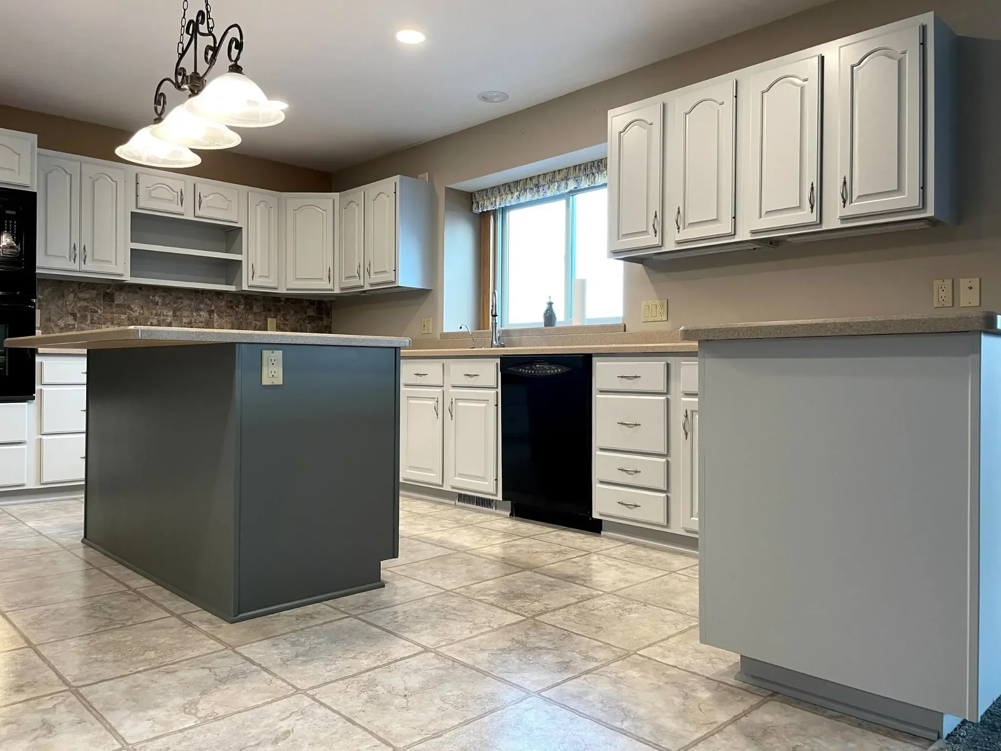 A kitchen with white cabinets, beige countertops, a dark gray island, a tiled backsplash, a large window with a valance, and tiled flooring. Overhead lighting fixture with three globe lights.